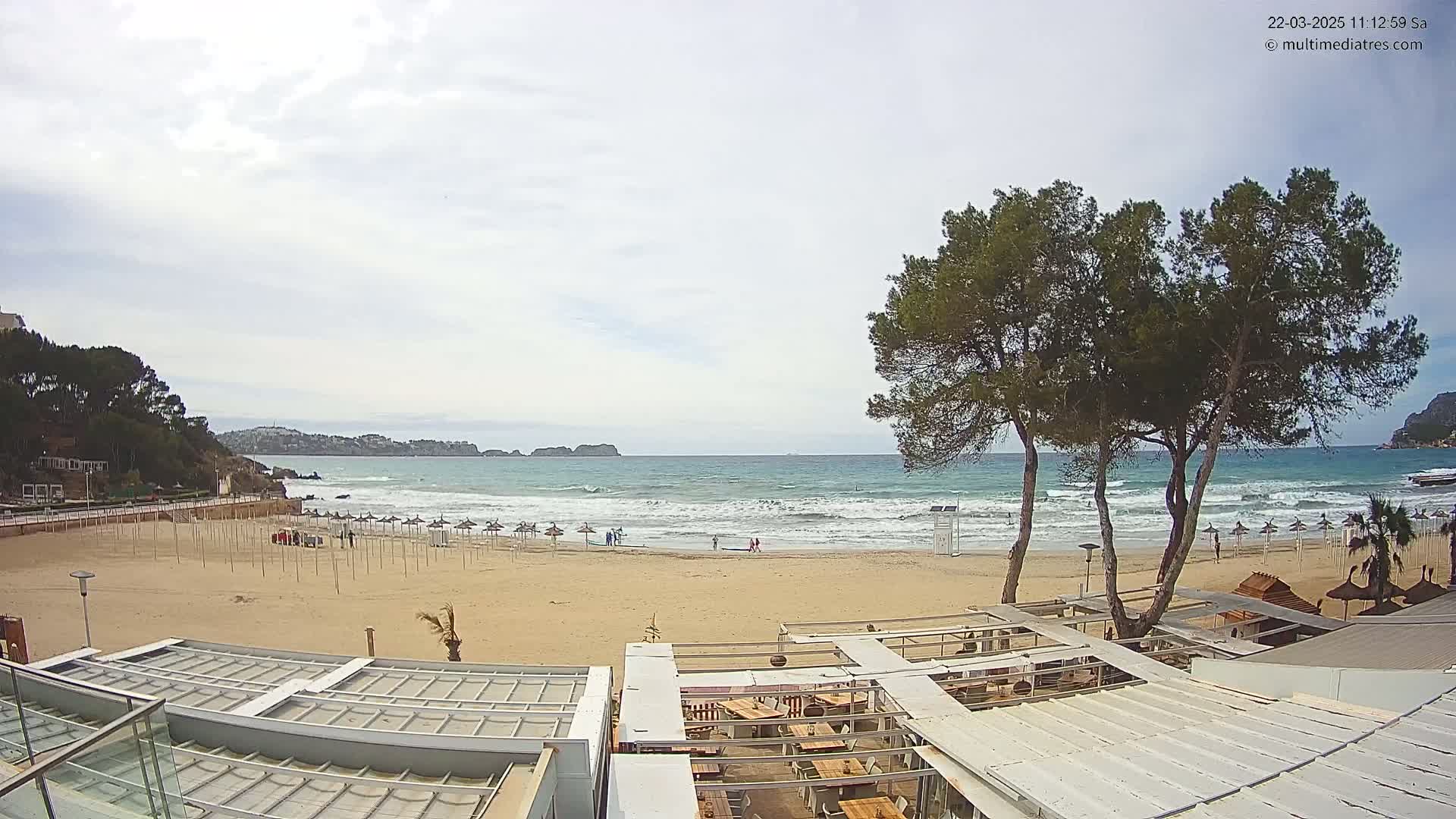 A mostly sunny day at a beach shows several closed umbrellas on the sand, a few people walking, a calm sea with small waves, and trees in the foreground next to an outdoor restaurant.