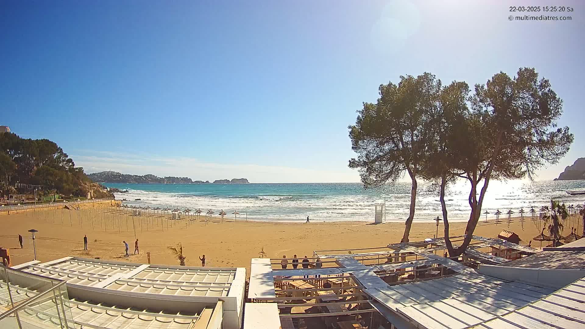 A sandy beach with calm waves under a clear blue sky, sparsely populated with people and umbrellas, is partially obscured by the roofs of a beachfront building and several pine trees.