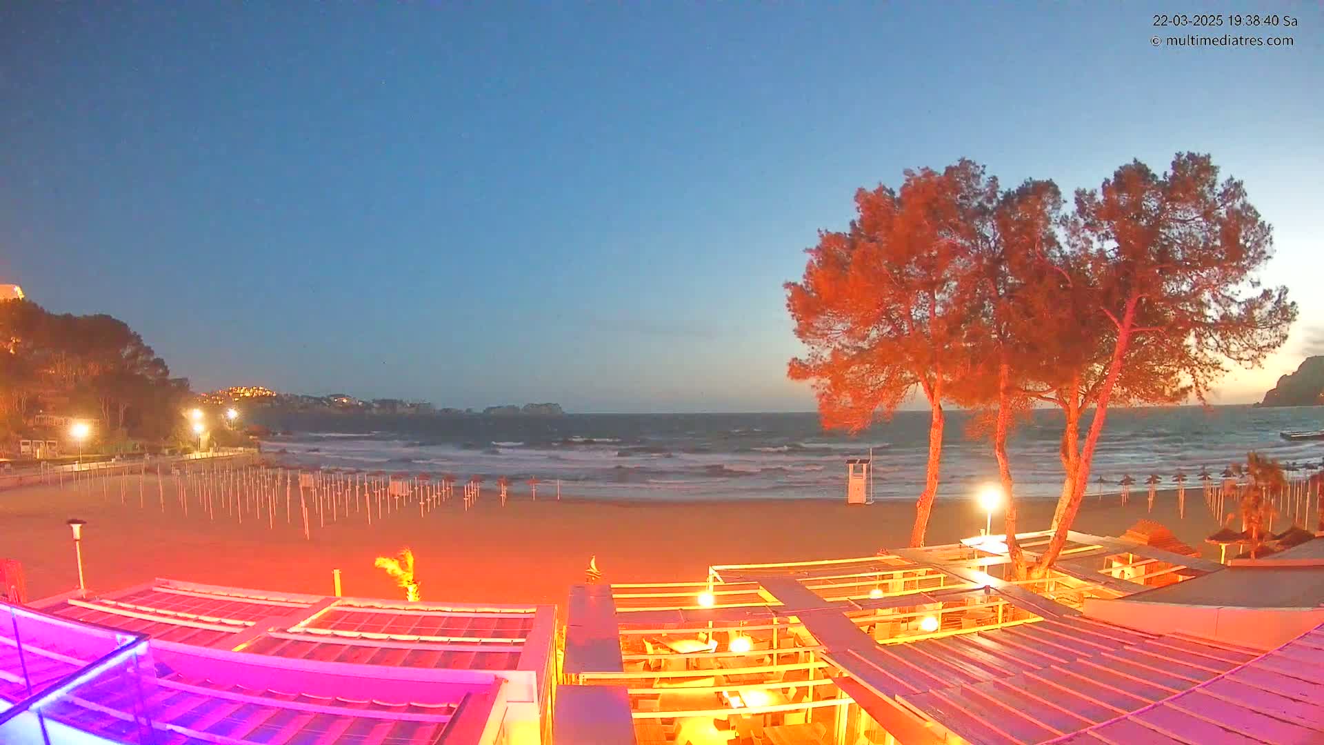 A beach at twilight with a somewhat choppy sea, illuminated beach umbrellas, and a brightly lit beachfront restaurant under a clear, dark-blue sky.