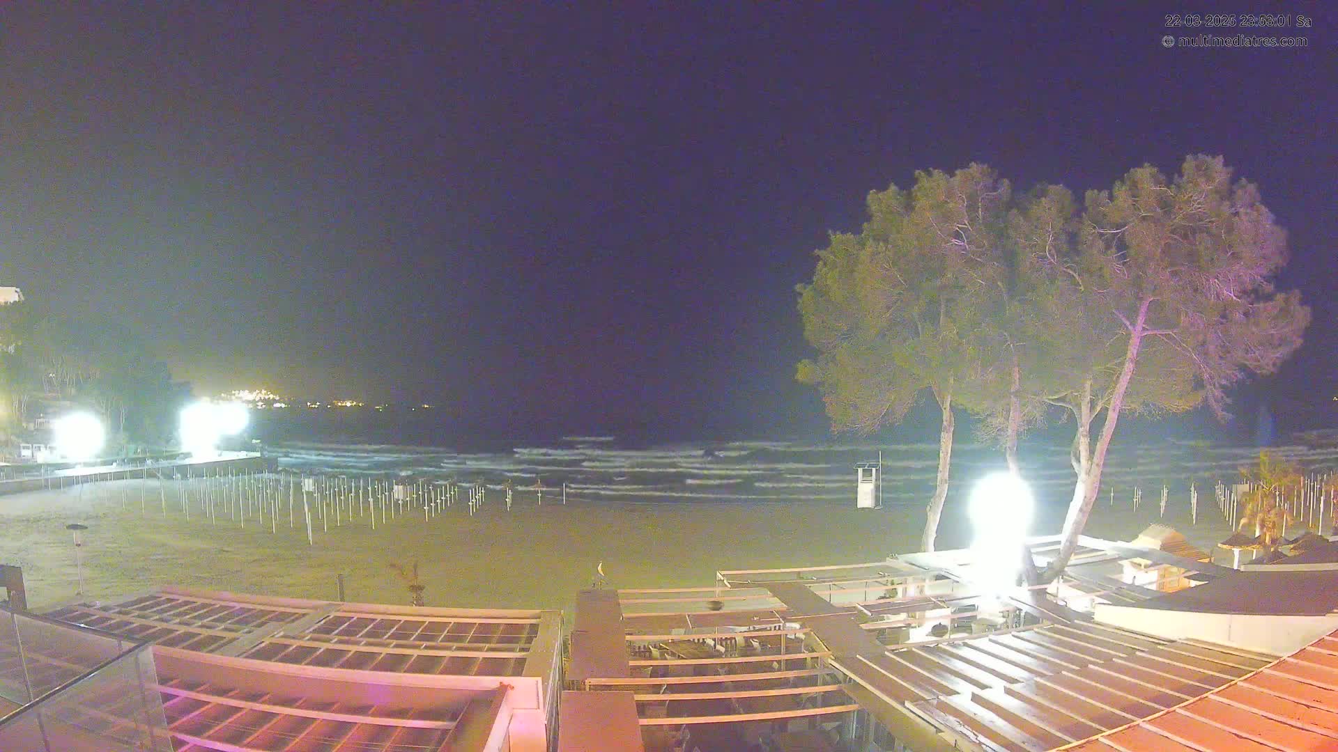 A nighttime view of a beach with gentle waves, illuminated by artificial lights, showing rows of beach umbrella stands and several structures along the shoreline under a clear, dark sky.