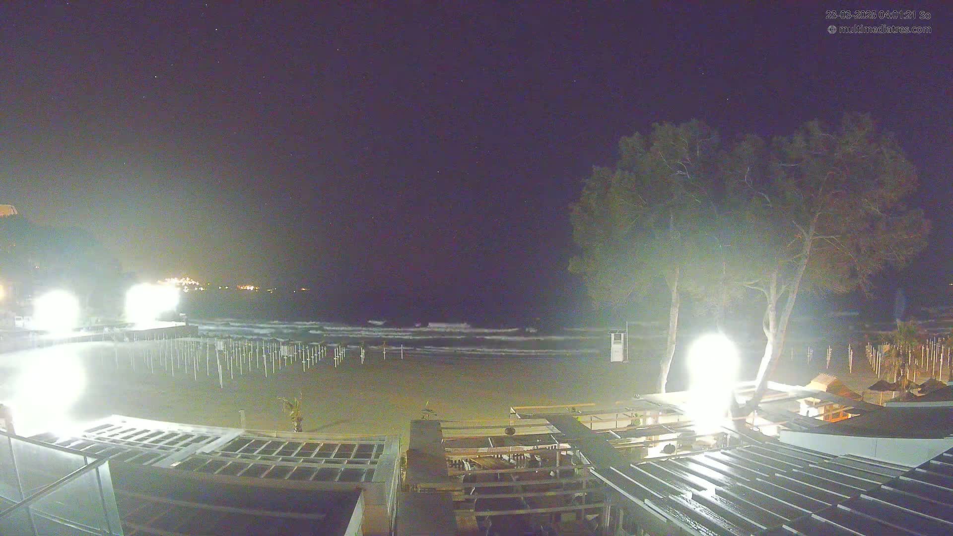 A nighttime beach scene under a clear, starry sky shows gently waving water, empty beach chairs, and brightly lit beachside structures.