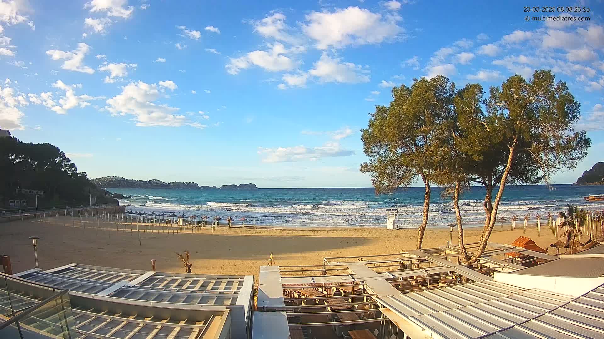 A sandy beach with gentle waves under a partly cloudy blue sky, viewed from above a beachside restaurant, with several pine trees in the foreground.