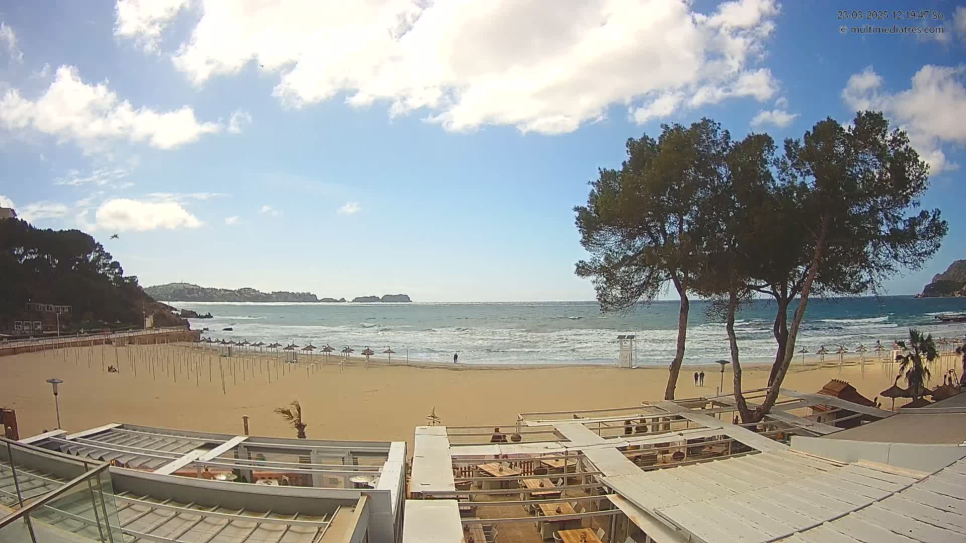 A mostly sunny day at a sandy beach with moderate waves, showing closed beach umbrellas and a beachfront restaurant with outdoor seating.