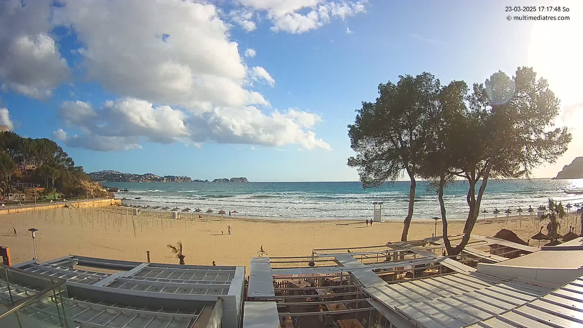 A mostly sunny day at a sandy beach shows a calm ocean with small waves, several people on the beach, and beach umbrellas lined up near the water's edge; some buildings and trees are in the foreground.