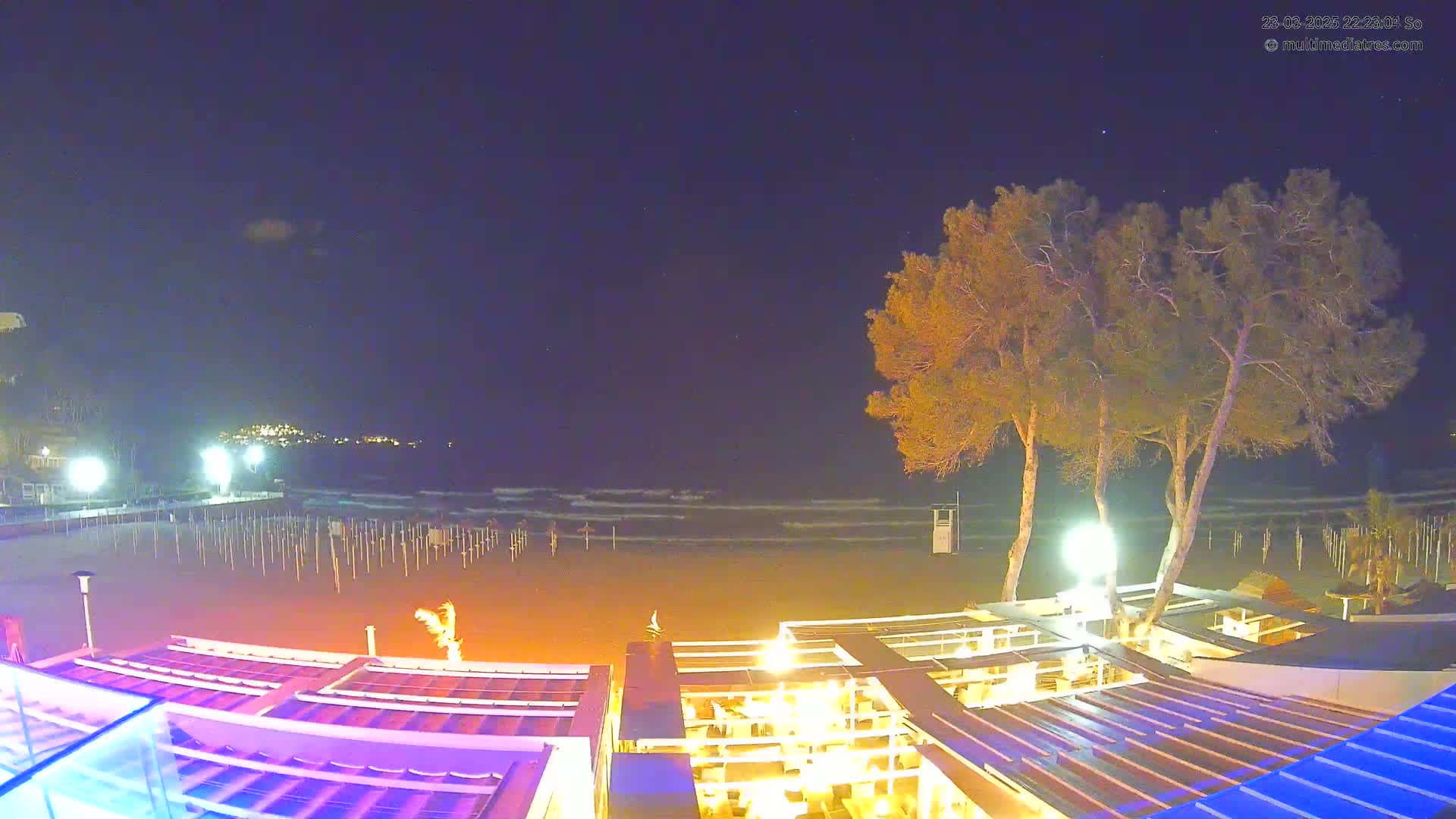 A nighttime beach scene shows a mostly empty sandy beach with gentle waves, illuminated beach umbrellas, and a brightly lit outdoor restaurant area under a clear, starry sky.