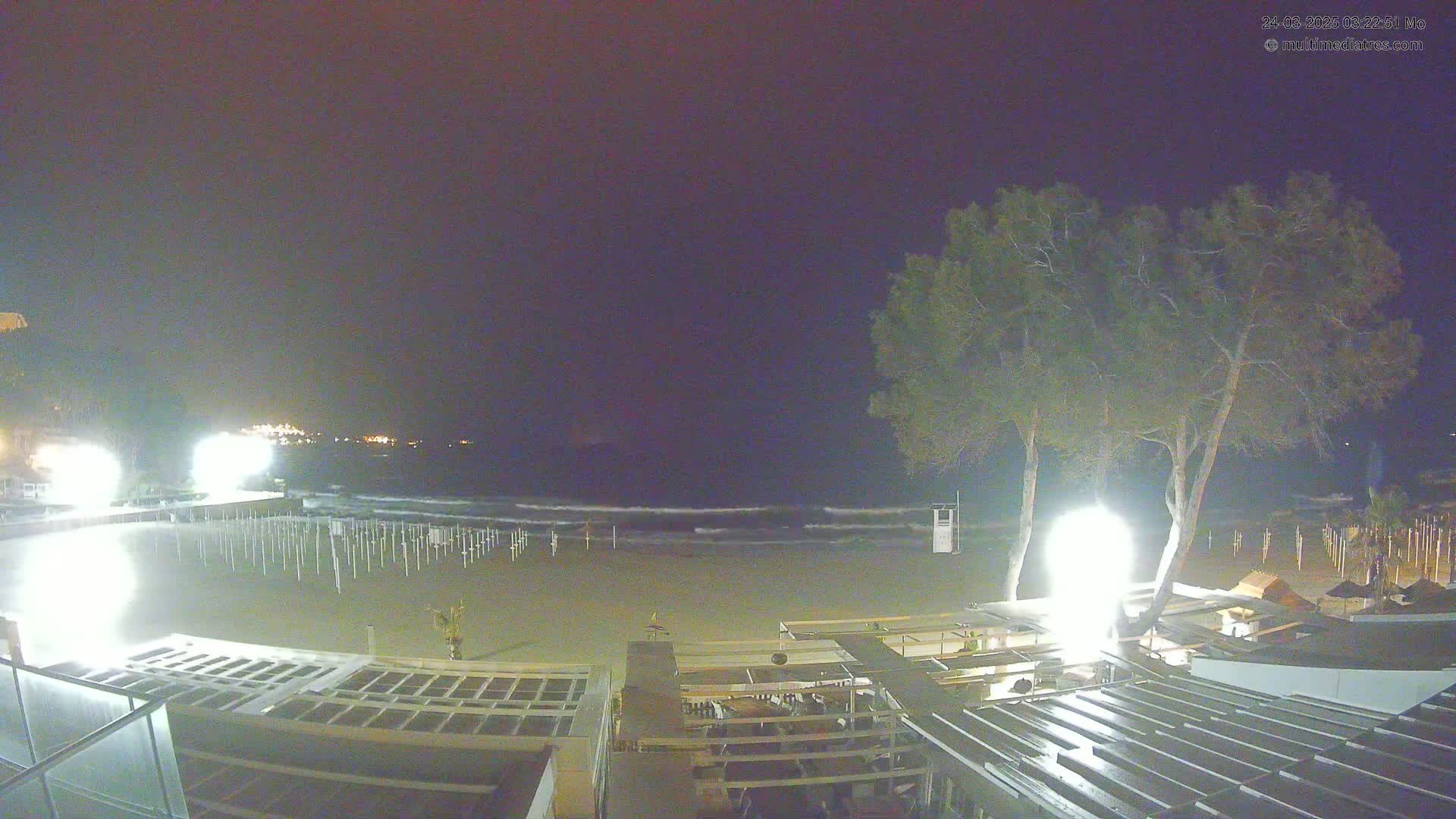 A nighttime view of a beach with calm waves under a dark sky, illuminated by artificial lights from beachside structures and trees.