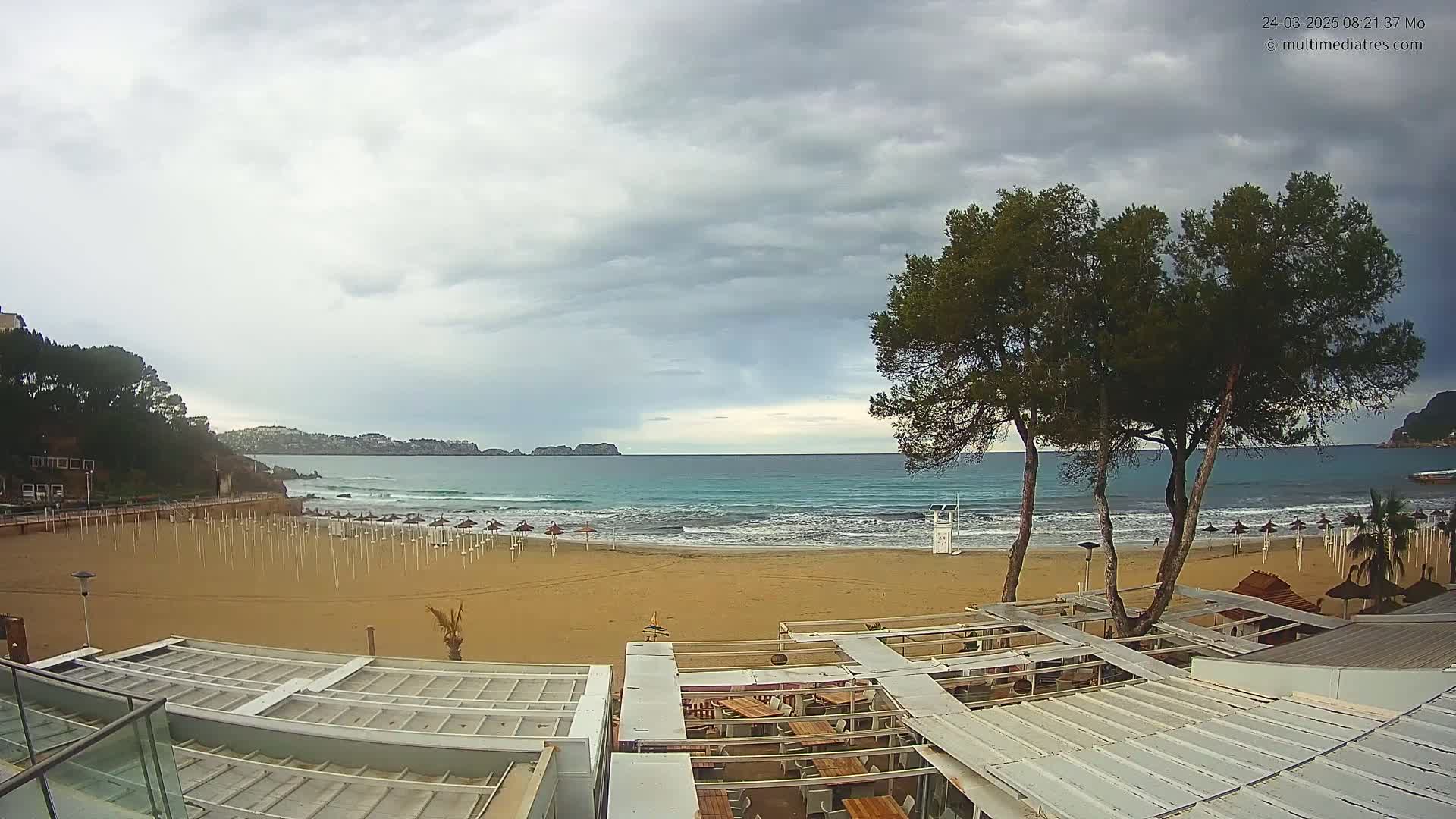 A mostly cloudy day at a beach shows a sandy shore with closed umbrellas, a lifeguard stand, and ocean waves lapping the shore, with trees and buildings in the foreground.