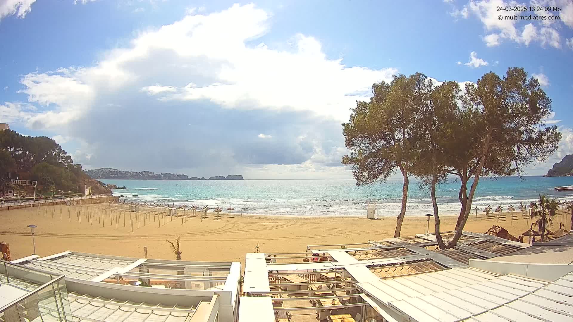 A sandy beach with rows of closed beach umbrellas and a partially-covered outdoor restaurant is seen under a partly cloudy sky, overlooking a calm, turquoise ocean.