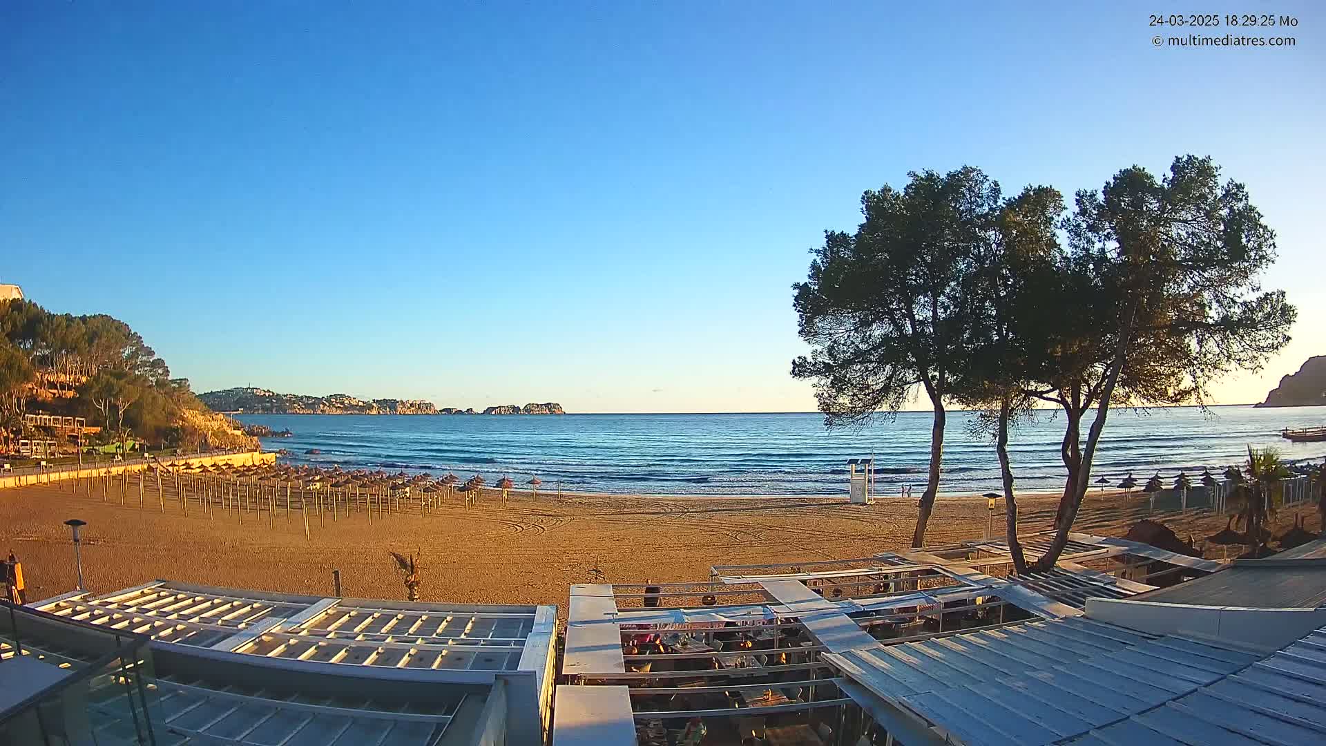 A sandy beach with closed umbrellas and a calm ocean under a clear blue sky is viewed from a beachside structure.