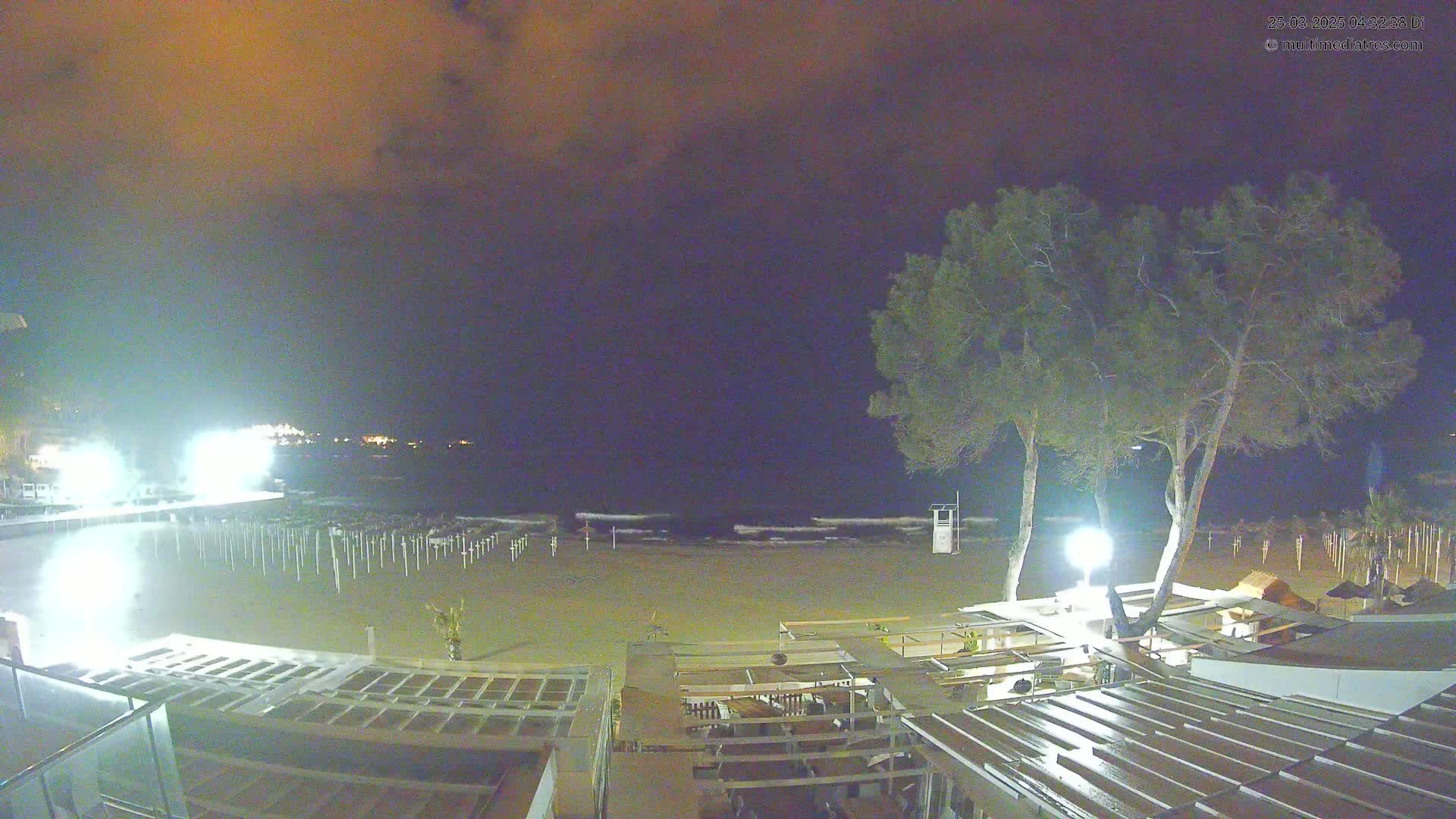 A nighttime view of a sandy beach with calm waves under a cloudy, dark orange sky, showing beach umbrellas folded down and two pine trees near beachside structures.