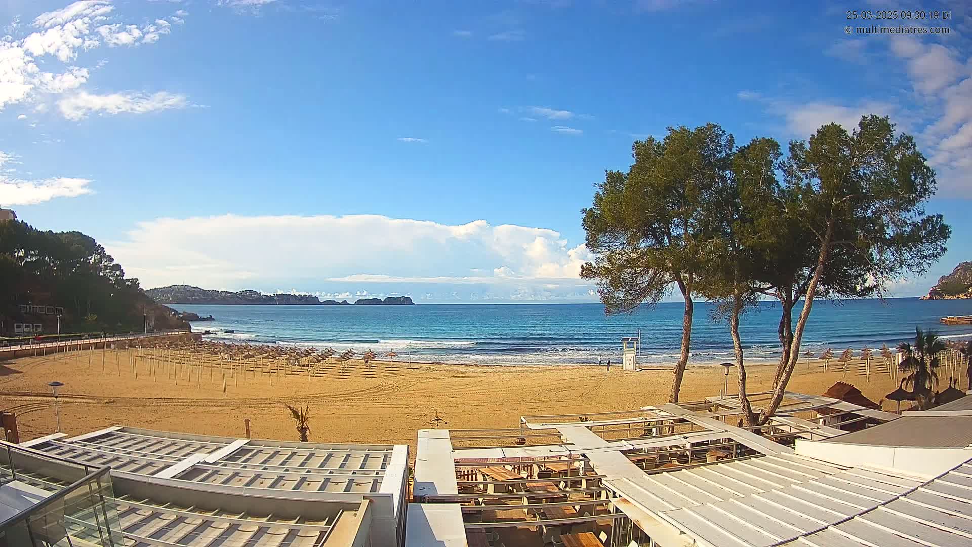 A sandy beach with rows of closed umbrellas and beachside structures is shown under a mostly sunny sky with some clouds, overlooking a calm ocean with small waves.