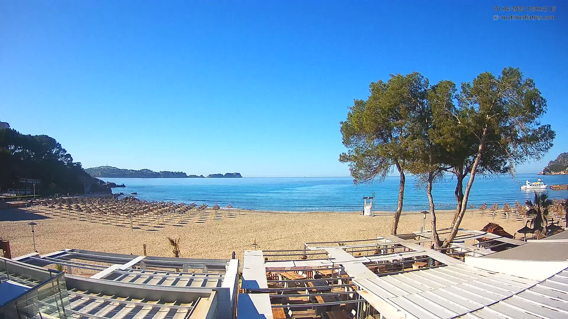 A sandy beach with numerous closed umbrellas stretches toward a calm, blue sea under a clear, sunny sky; several trees and a partially visible building are in the foreground.