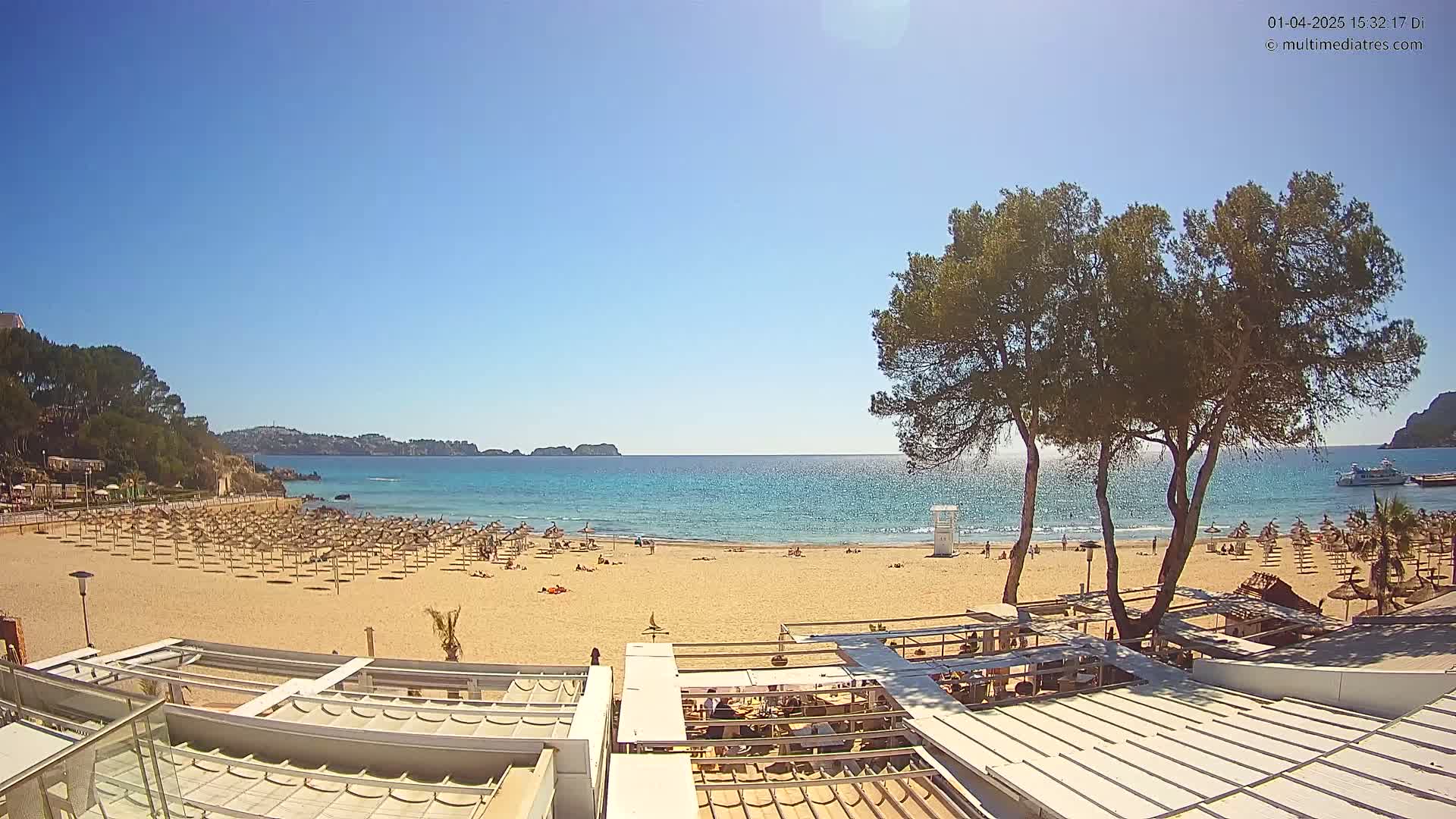A sandy beach with numerous umbrellas and sunbathers is visible, overlooking a calm, clear blue ocean under a bright sunny sky, with trees and a beachfront building in the foreground.