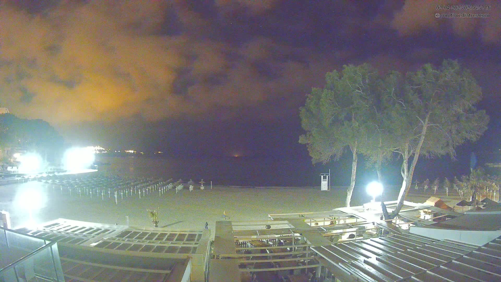 A nighttime view of an empty beach with closed umbrellas and beachside structures under a cloudy, dark sky.