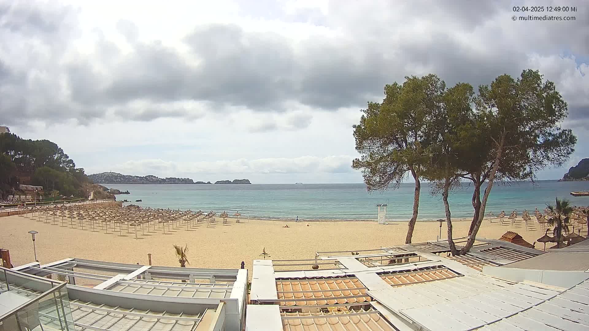 A mostly sunny beach with numerous closed umbrellas, a calm sea, and several pine trees is viewed from a high vantage point overlooking beachside buildings.