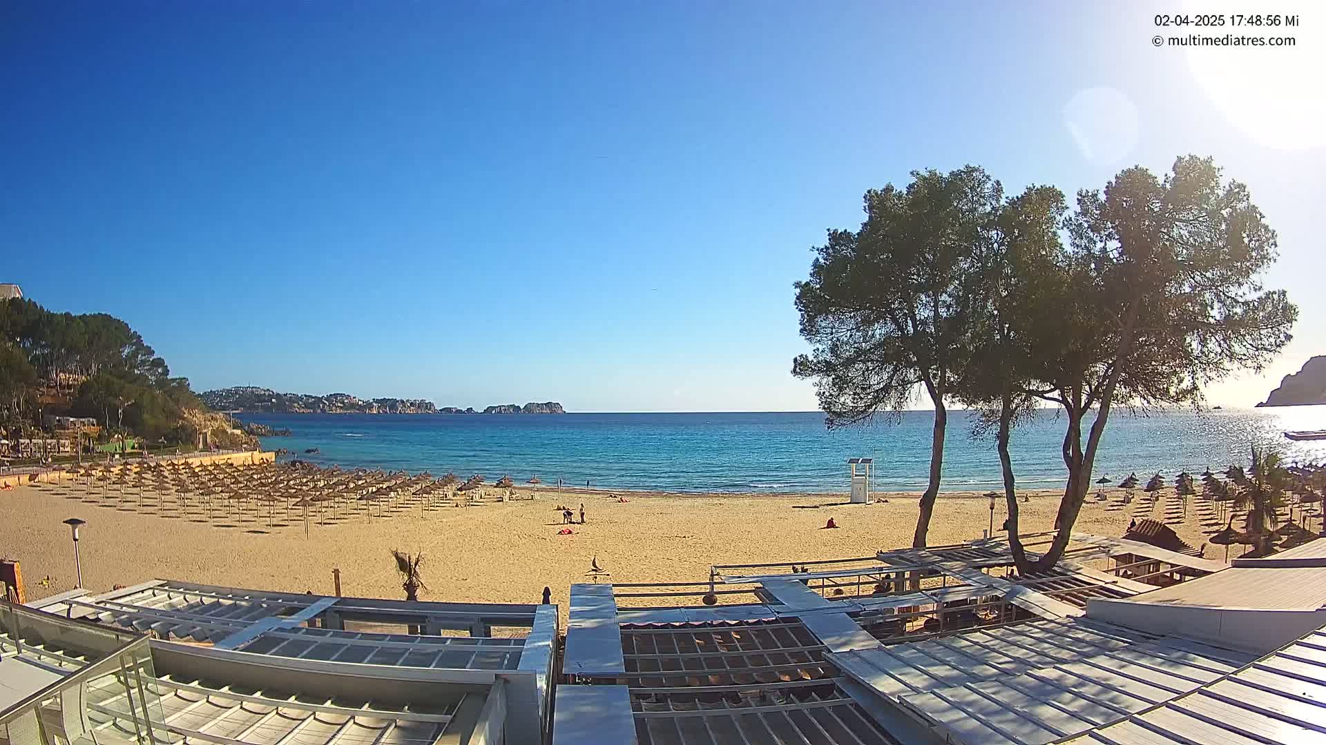 A sandy beach with numerous umbrellas and a few people is seen under a clear blue sky, with calm turquoise water and distant hills in the background.