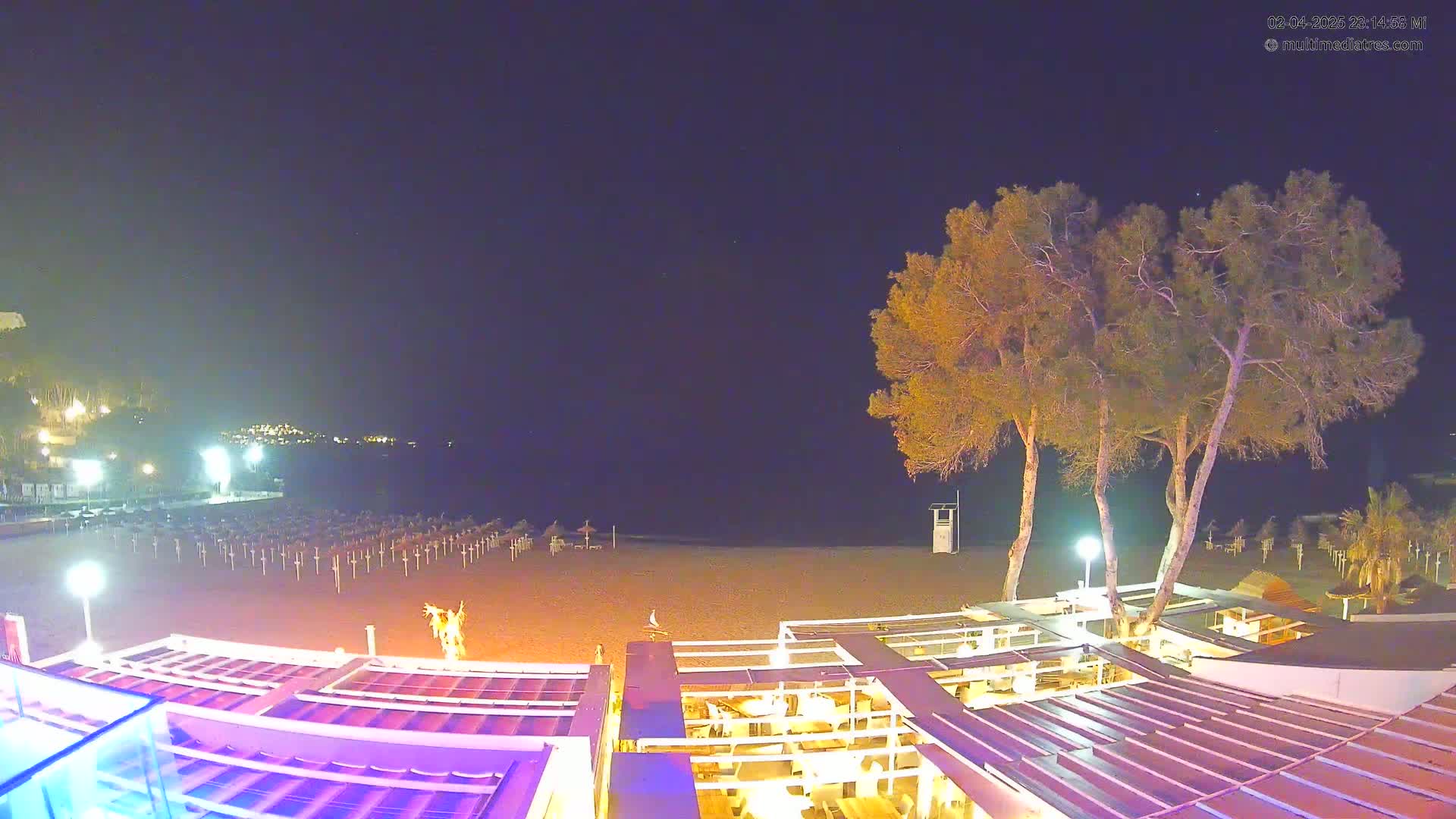 A nighttime beach scene shows a sandy shore with closed umbrellas, a few illuminated trees, and a beachfront restaurant with outdoor seating under a partially covered structure.