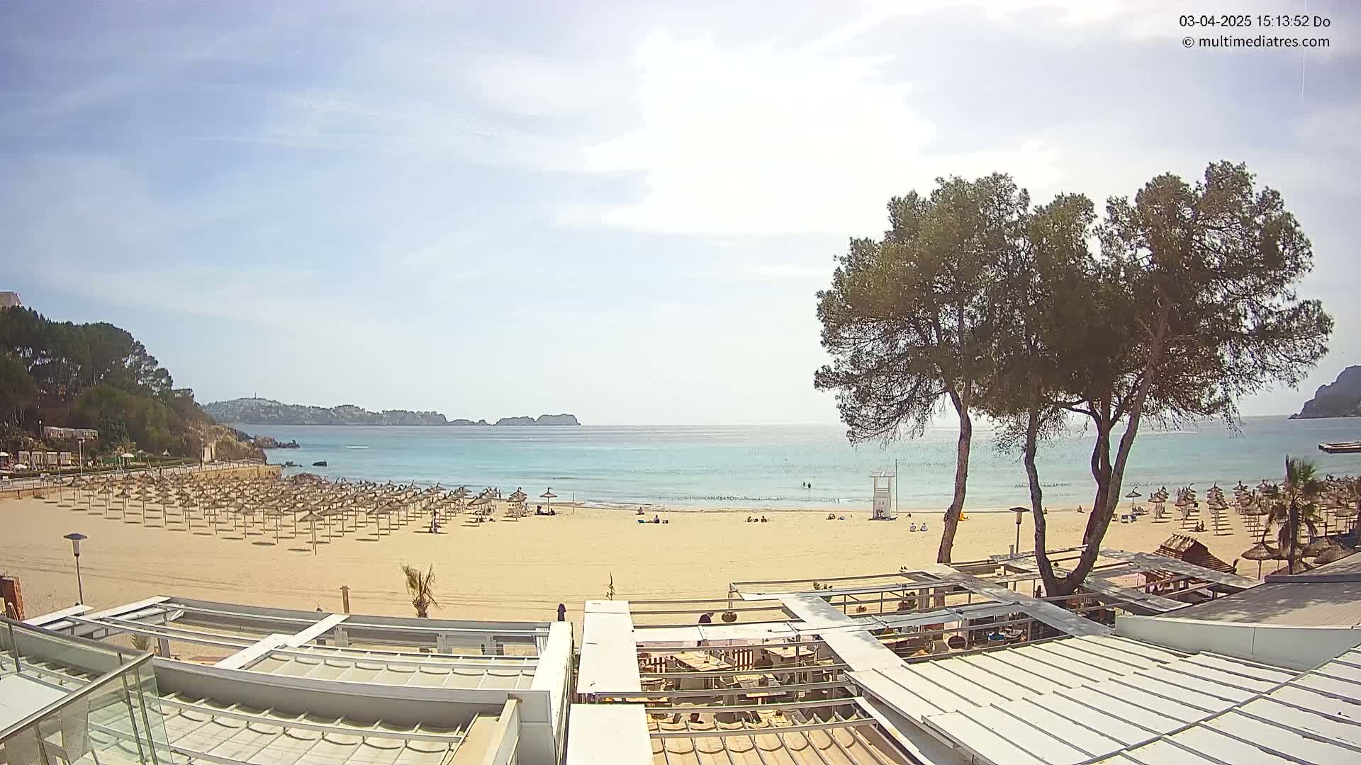 A sandy beach with numerous umbrellas and a few people is seen from an elevated vantage point on a sunny day, overlooking calm turquoise water and a distant shoreline.
