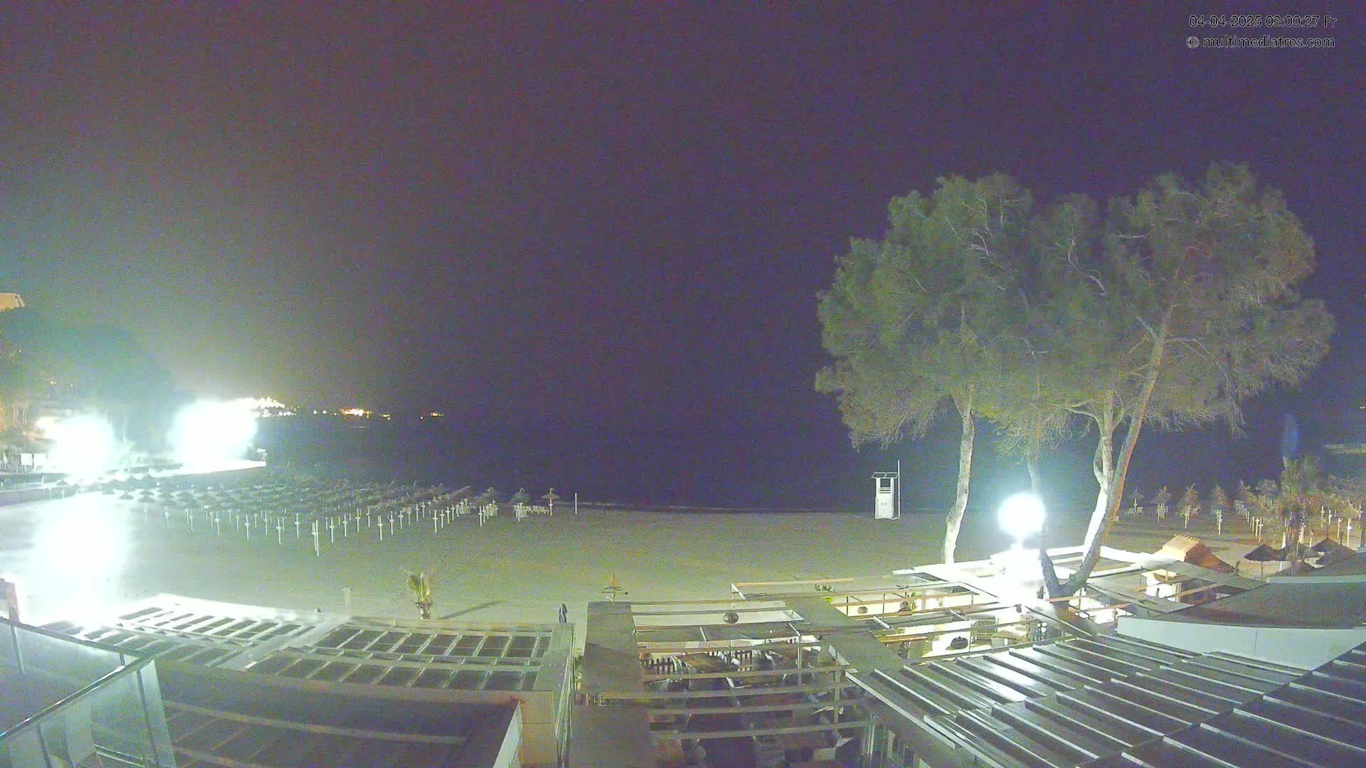A nighttime view of an empty beach with rows of closed beach umbrellas, a few trees, and outdoor seating areas under a dark sky.