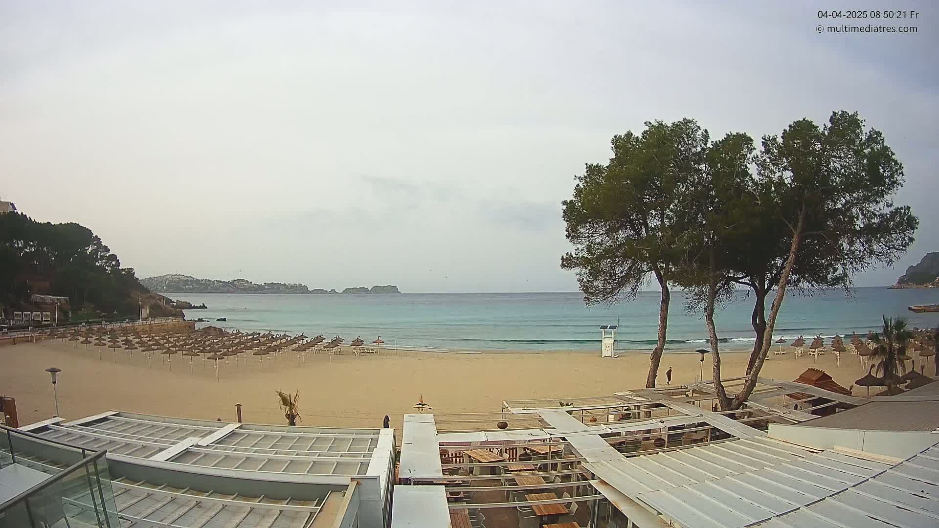A mostly clear, overcast day shows a sandy beach with rows of closed umbrellas and a beachfront restaurant, overlooking a calm, turquoise ocean with small waves.