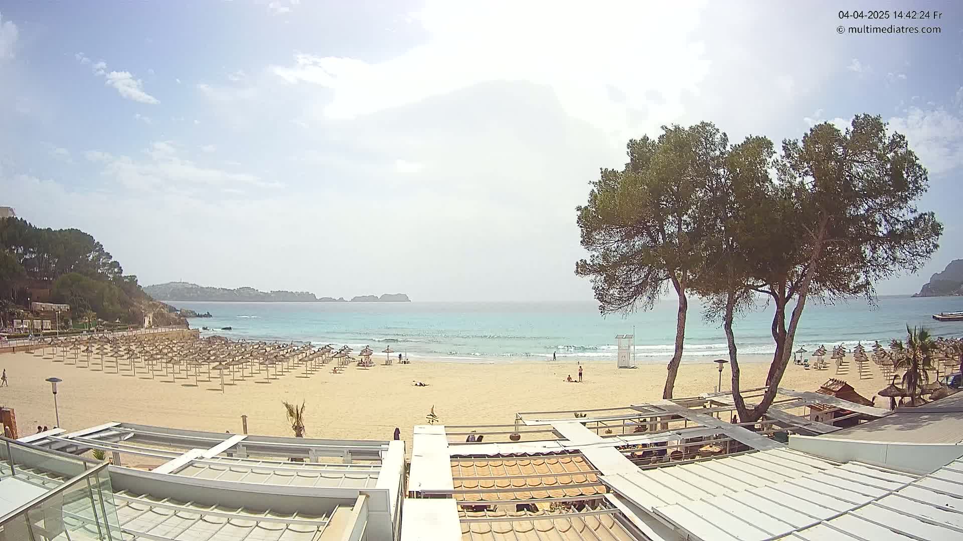 A sandy beach with rows of umbrellas and a calm turquoise ocean under a partly sunny sky is viewed from a rooftop patio.
