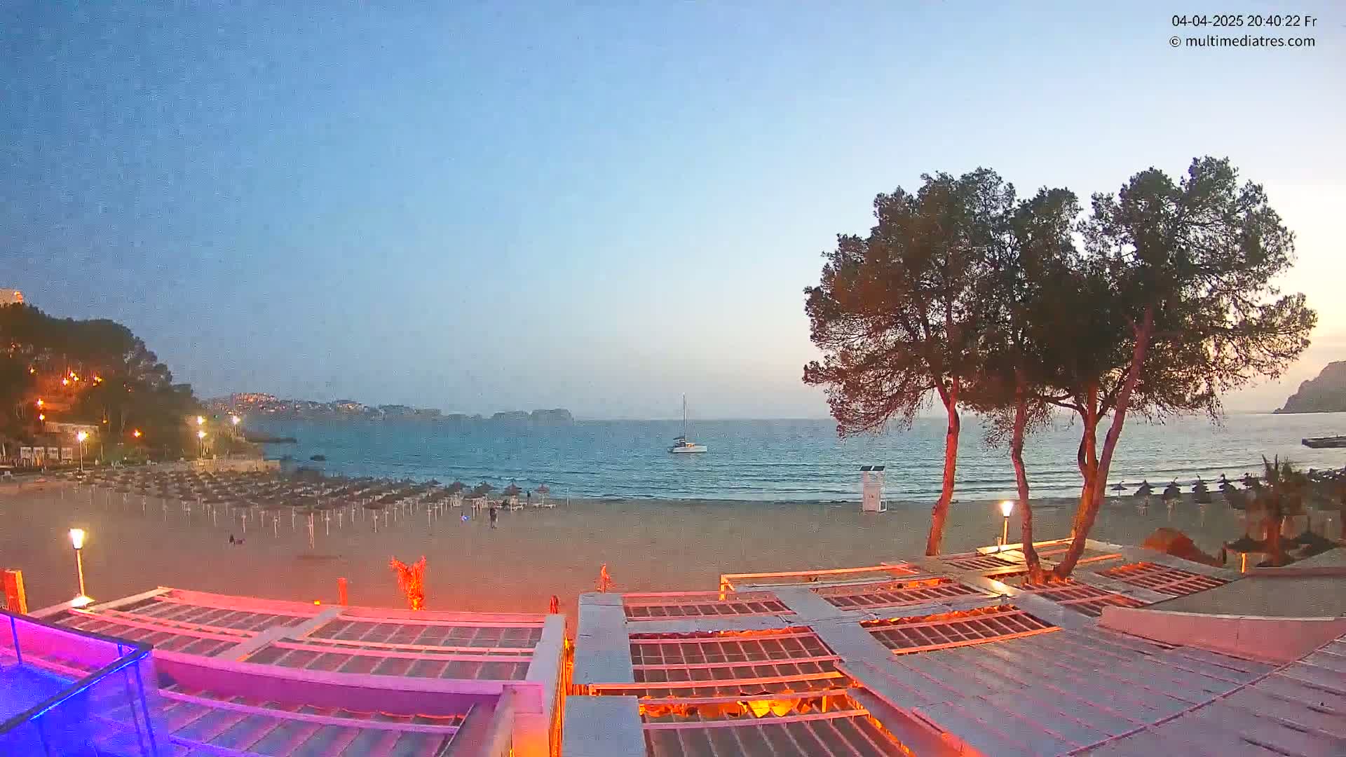 A sandy beach at twilight, with calm water, a sailboat on the horizon, and beach umbrellas folded for the night, is viewed from a beachfront establishment with illuminated rooftops.