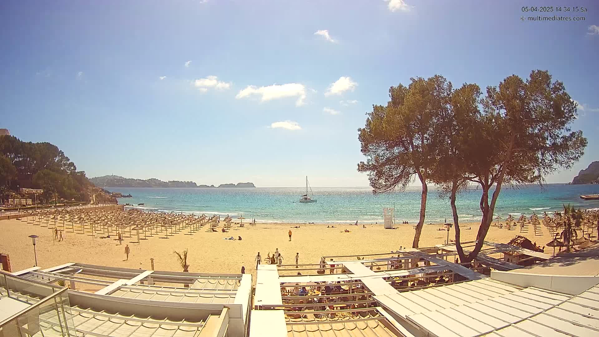 A sandy beach with numerous umbrellas and sunbathers is visible under a mostly sunny sky, with calm, turquoise water and a sailboat in the distance.