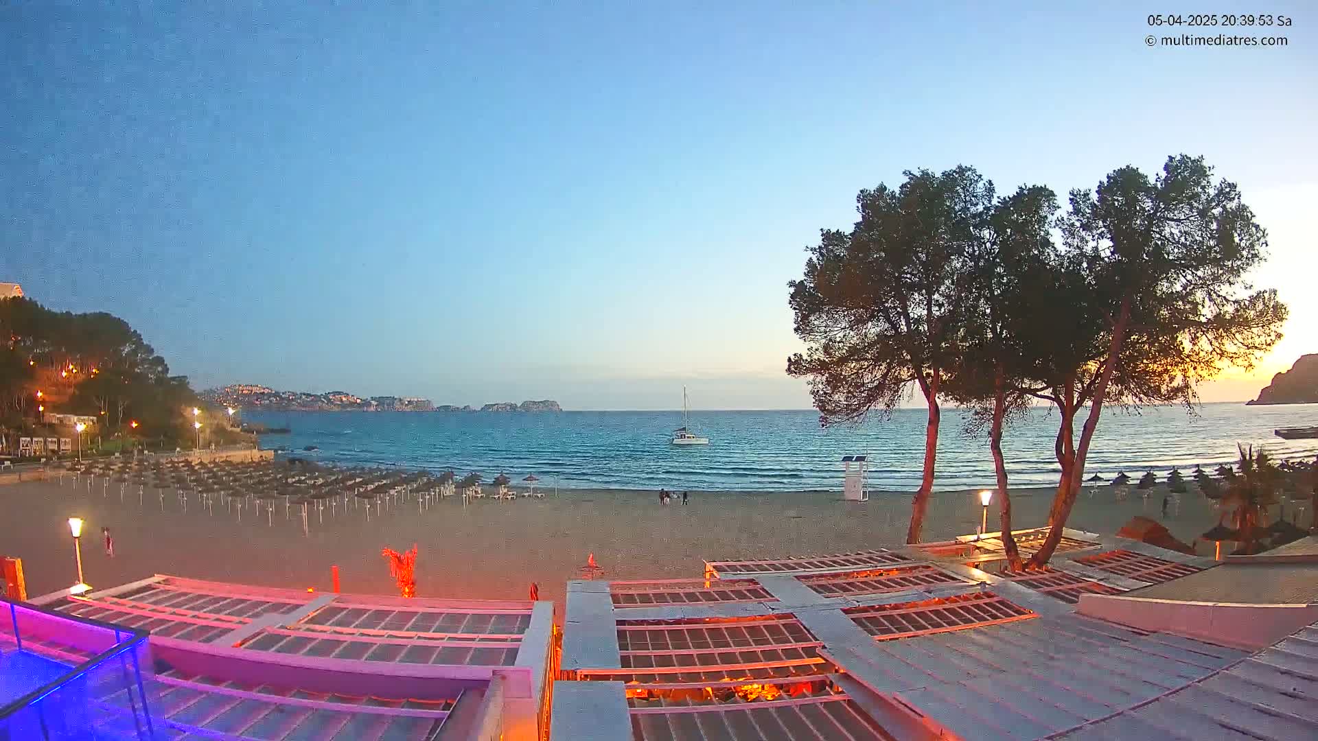 A sandy beach at dusk, with calm water, a few people, and several beach umbrellas, is viewed from above a partially lit outdoor structure, with trees in the foreground and hills in the distance.