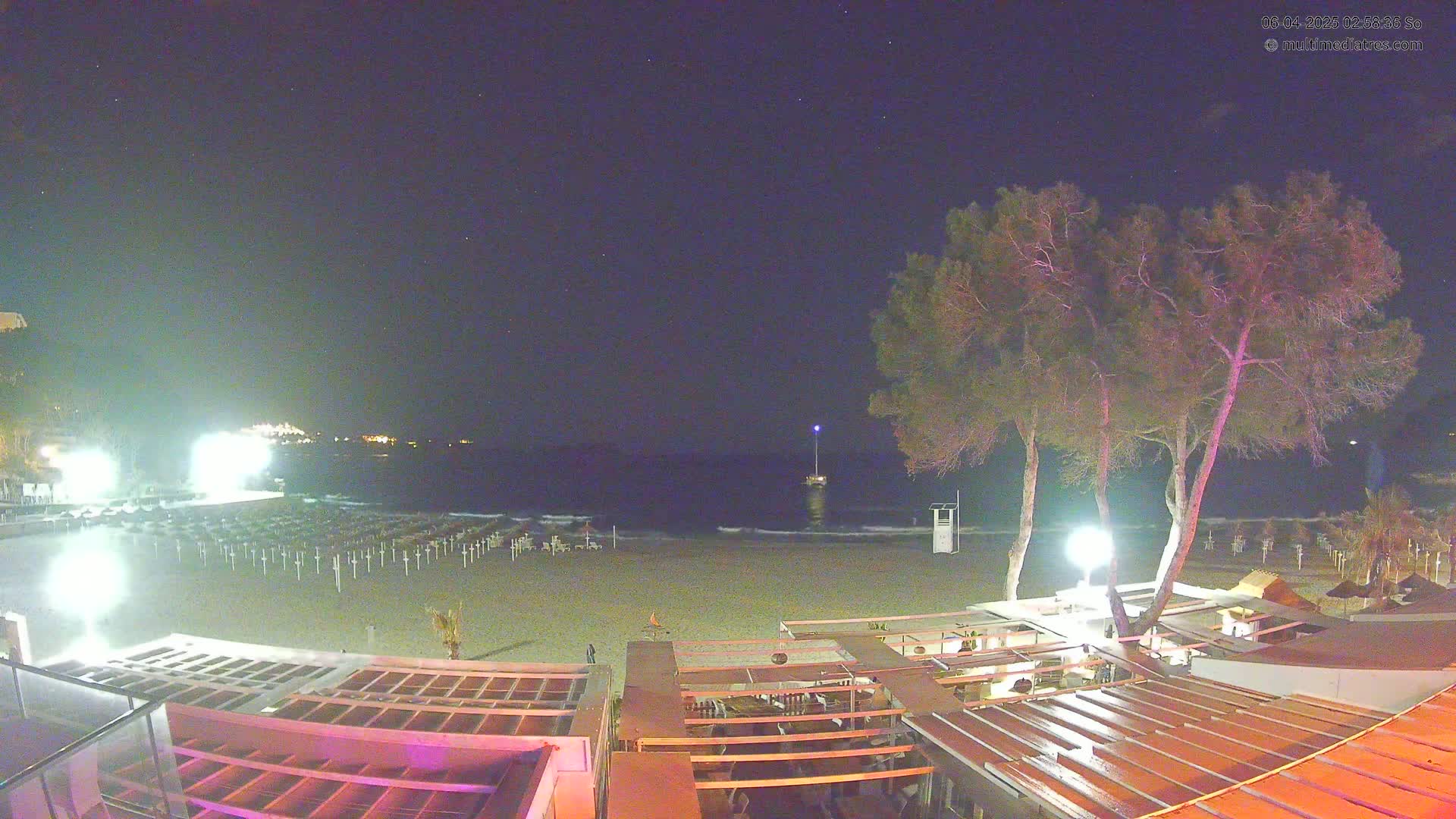 A nighttime view of a beach with closed umbrellas, a few lights, and several trees, overlooking a calm sea under a clear, starry sky.