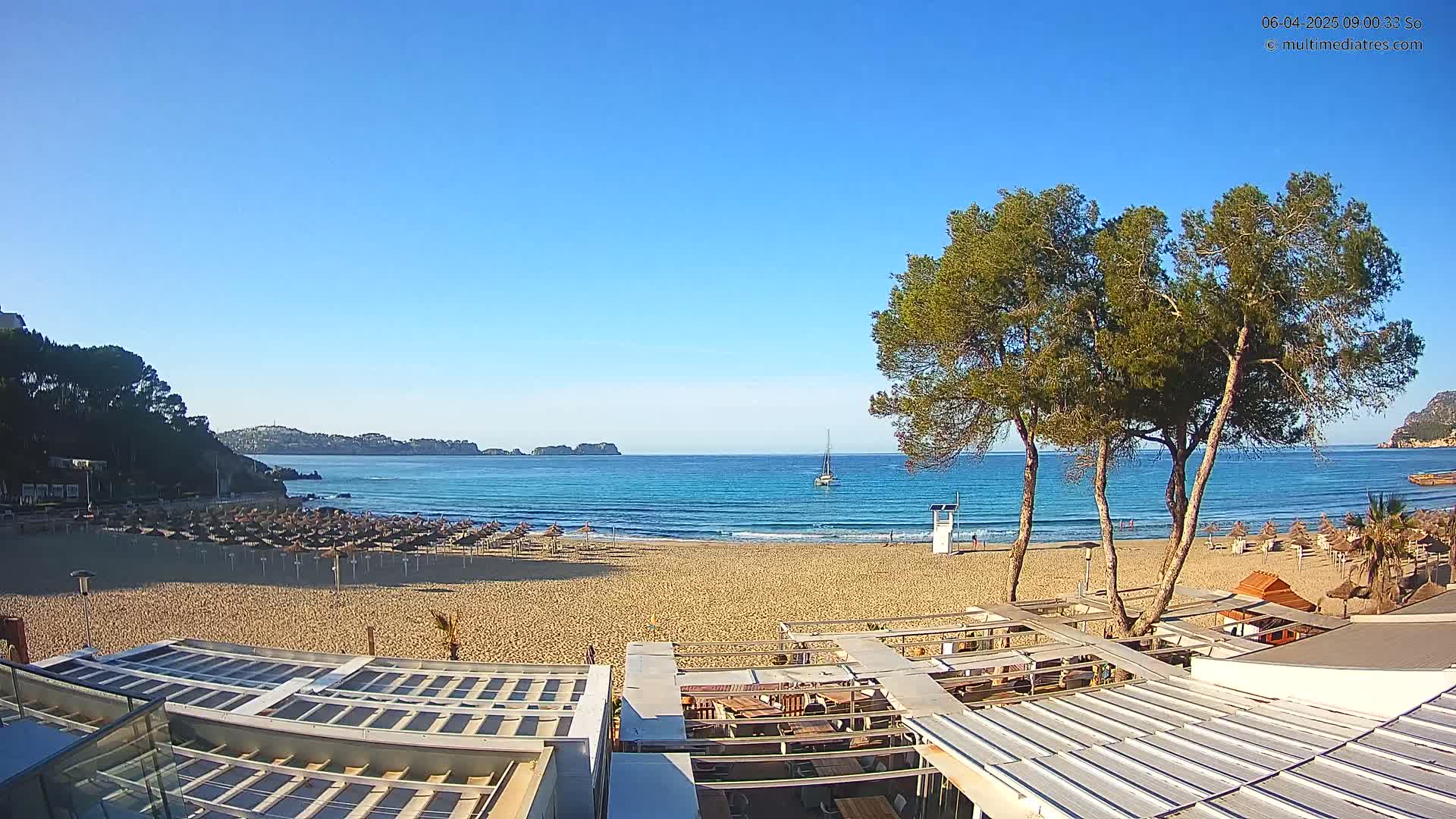 A sandy beach with rows of closed umbrellas and a calm, blue ocean under a clear, sunny sky is viewed from above a beachside structure.