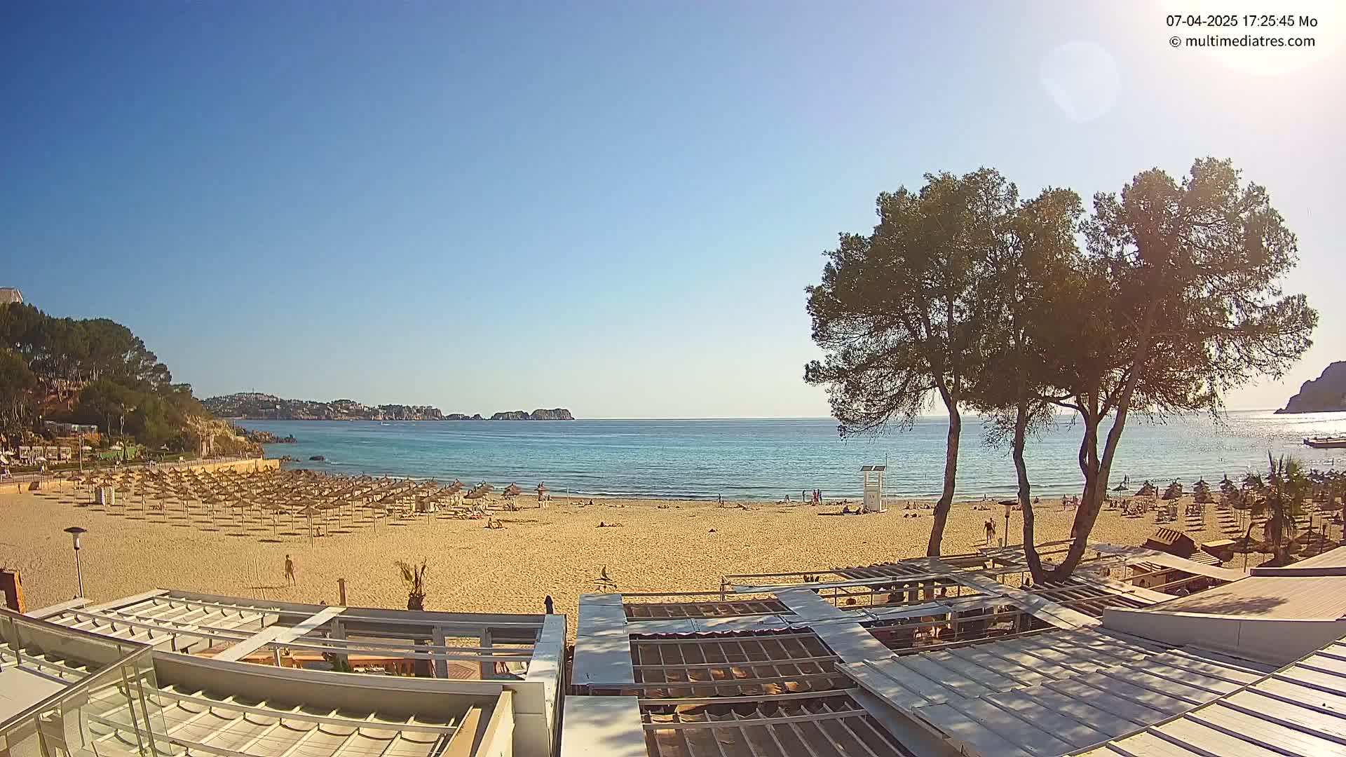 A sandy beach with numerous umbrellas and a few people, backed by trees and buildings, overlooks a calm, clear ocean under a bright, sunny sky.