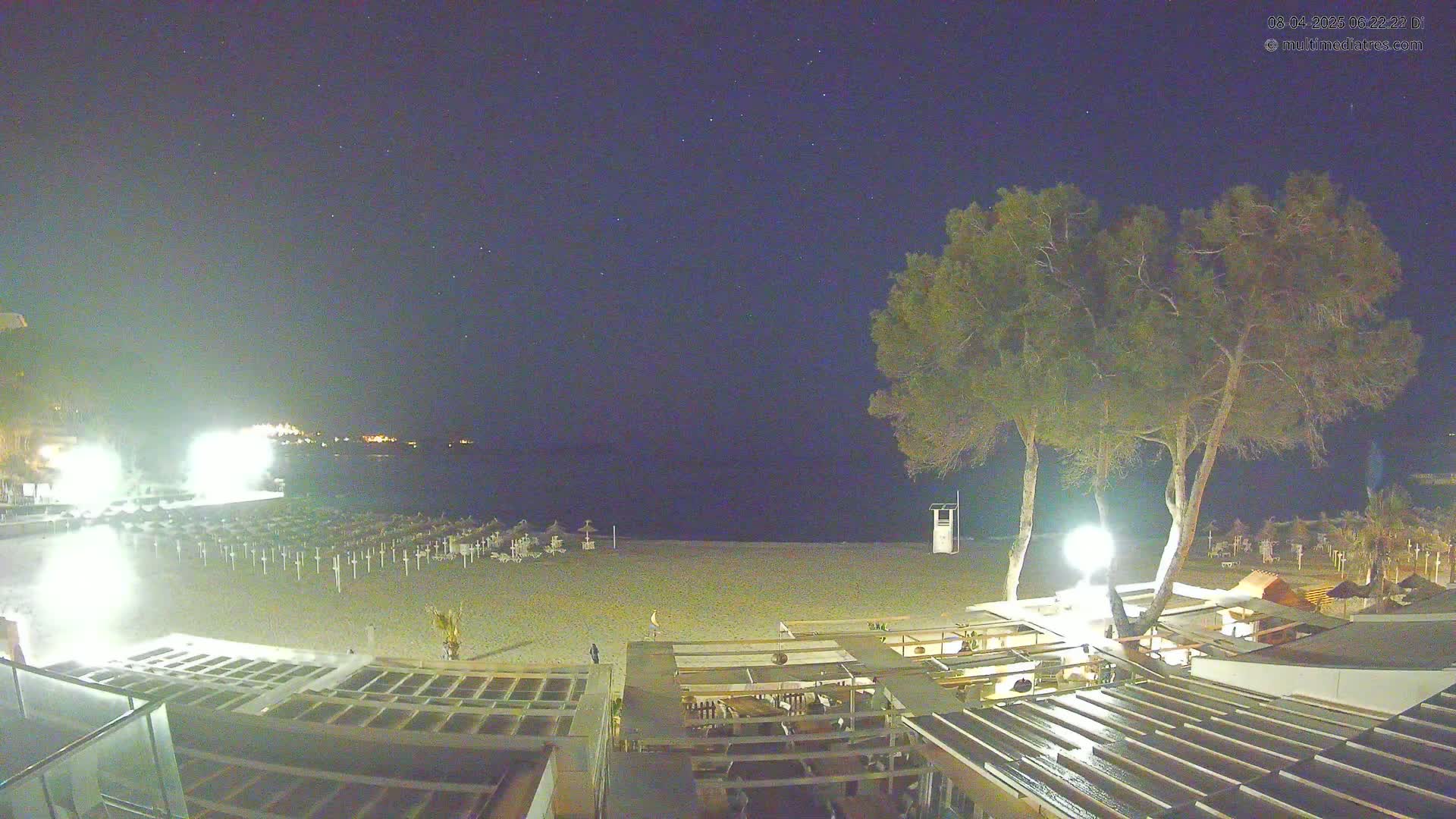 A nighttime beach scene under a clear, starry sky shows a sandy beach with closed umbrellas and a few people near a beachfront bar and restaurant, illuminated by artificial light.