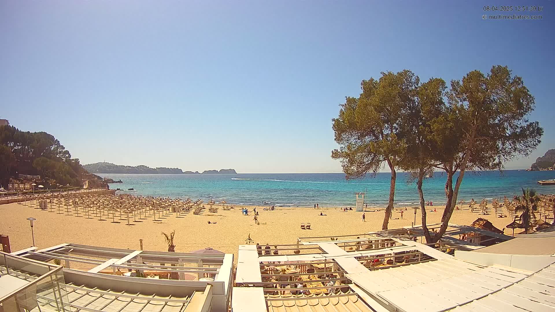 A sandy beach with numerous umbrellas and a few people, backed by a turquoise ocean and lush green trees under a clear blue sky.