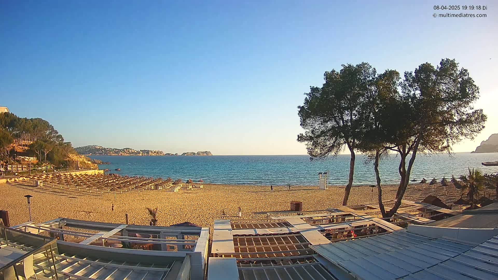 A sandy beach with rows of umbrellas and a calm sea under a clear blue sky is viewed from atop a building.