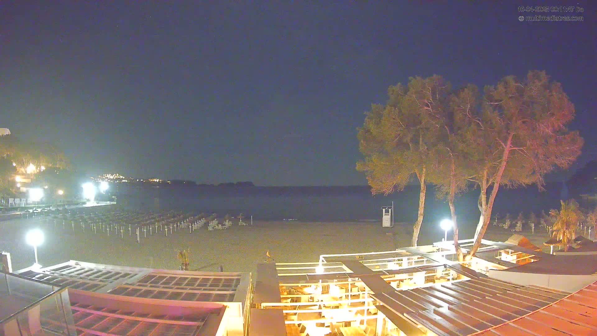 A nighttime beach scene shows a mostly empty sandy beach with rows of closed beach umbrellas, a calm dark sea, and a partially lit outdoor restaurant structure with trees nearby under a clear, dark sky.