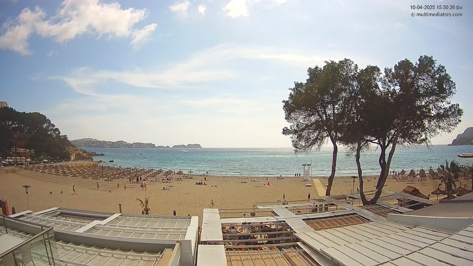 A mostly sunny day at a sandy beach with calm turquoise water, numerous beach umbrellas, and people enjoying the shore, observed from above a beachfront building.