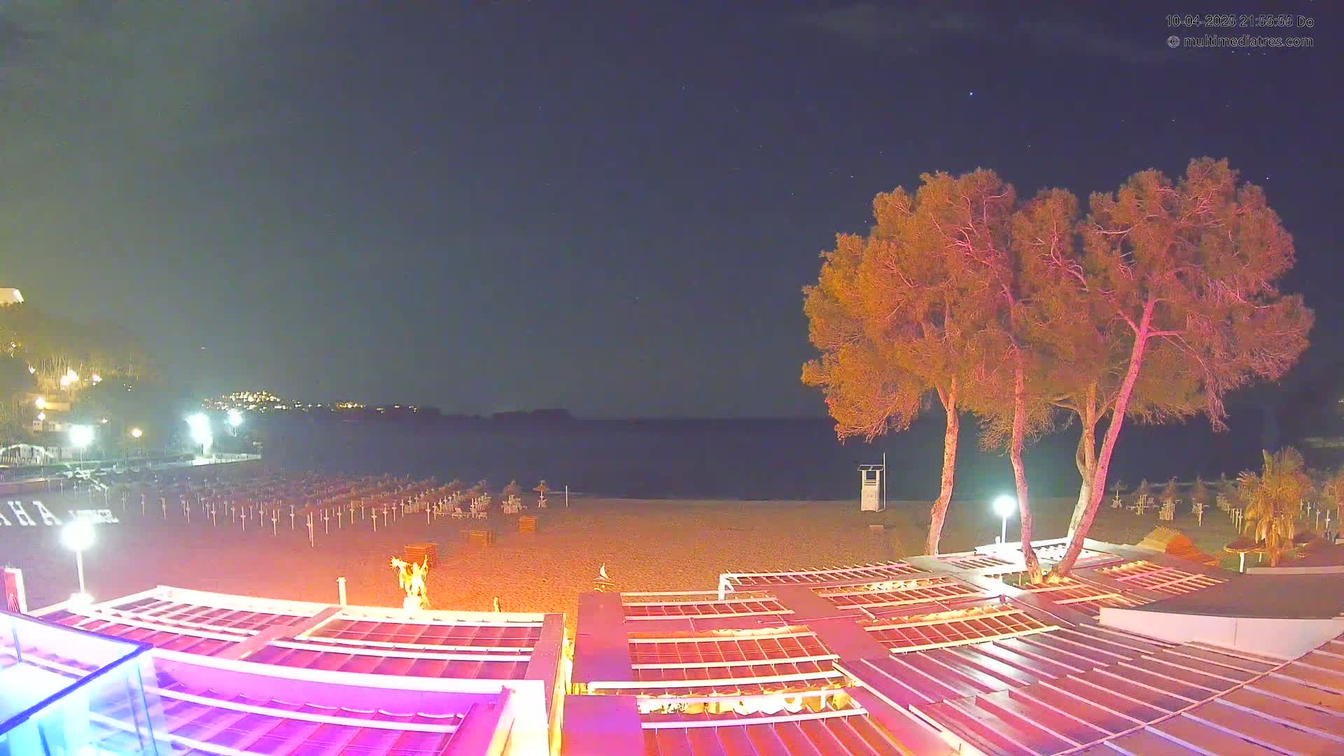 An empty beach at night under a clear, starry sky is illuminated by artificial lights, with several trees and beach umbrellas visible near a beachfront establishment.