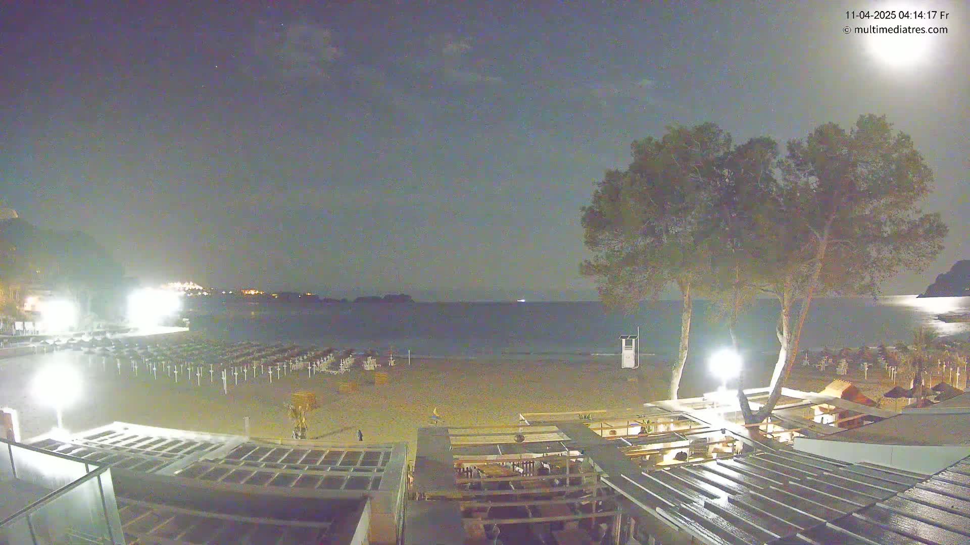 A nighttime view of a mostly empty beach with rows of closed beach umbrellas, illuminated by artificial lights, under a partly cloudy sky.