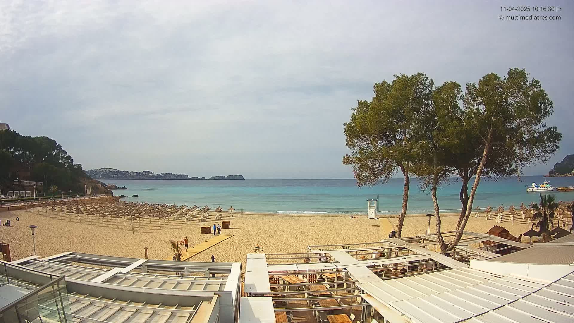 A sandy beach with rows of umbrellas and a calm, turquoise ocean under a partly cloudy sky is viewed from above a beachside restaurant.