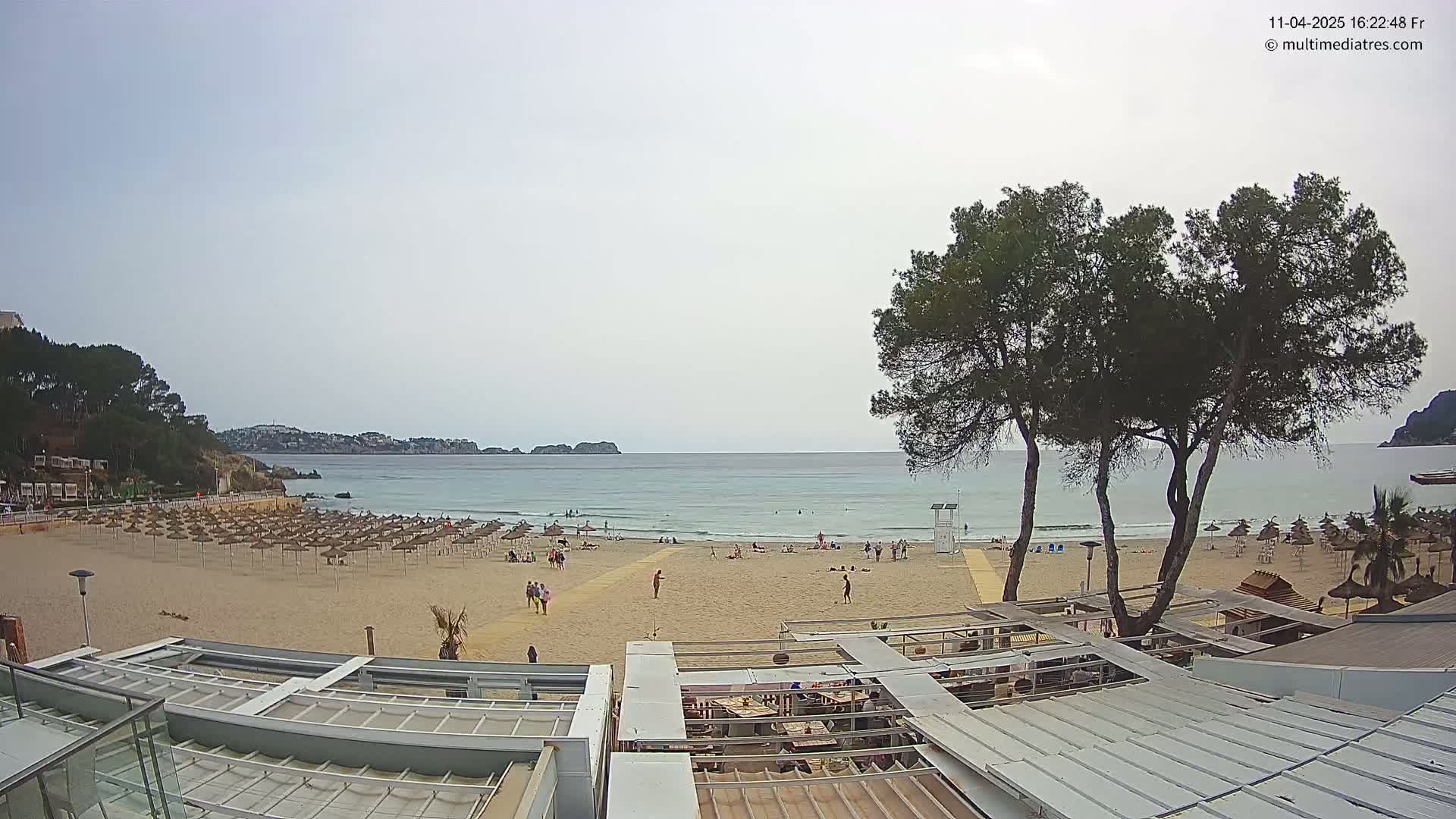 A sandy beach with numerous umbrellas and a few people is seen from above, overlooking a calm ocean under a mostly cloudy sky, with green trees in the foreground and buildings along the shoreline.