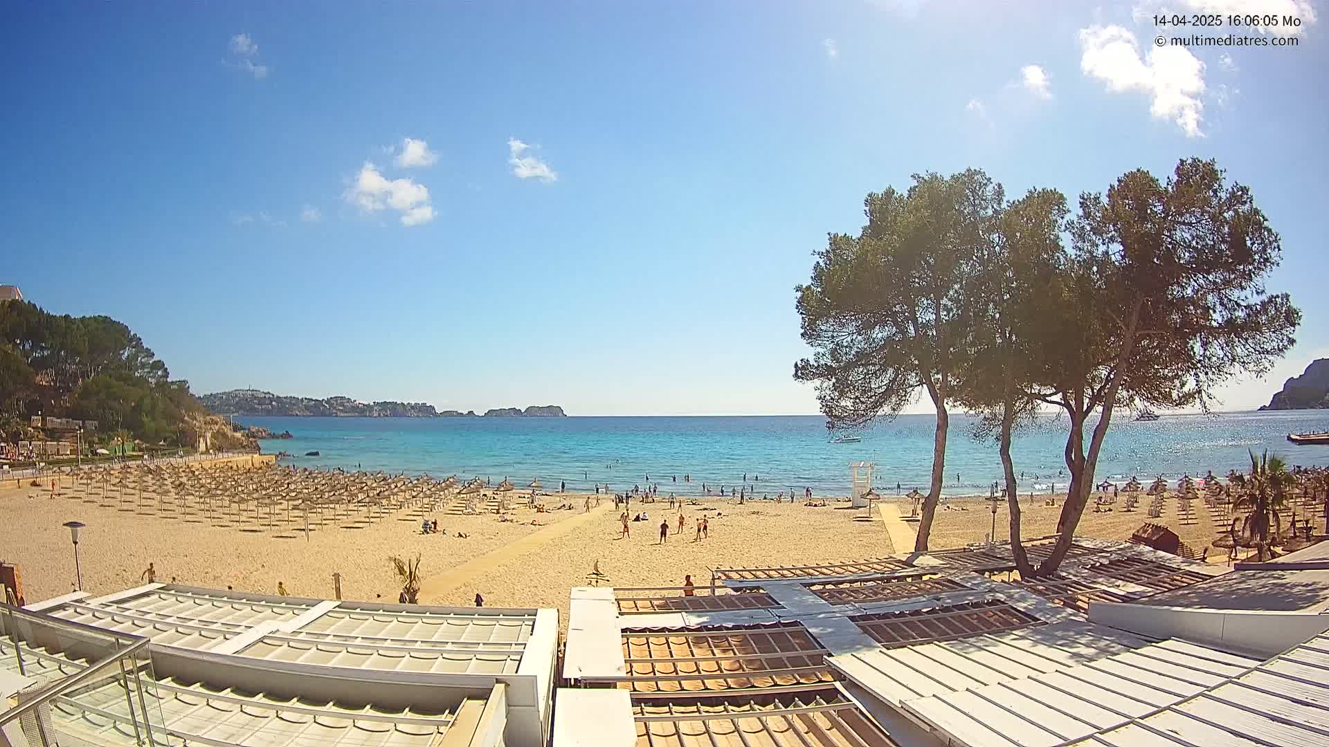 A sandy beach with numerous umbrellas and sunbathers is visible, under a clear blue sky with a few clouds, overlooking a calm, turquoise sea.