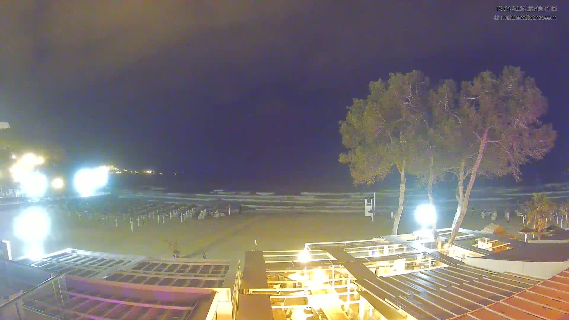 A nighttime beach scene shows gently lapping waves under a dark sky, illuminated beach umbrellas, and the lighted structures of a beachfront establishment near two tall trees.