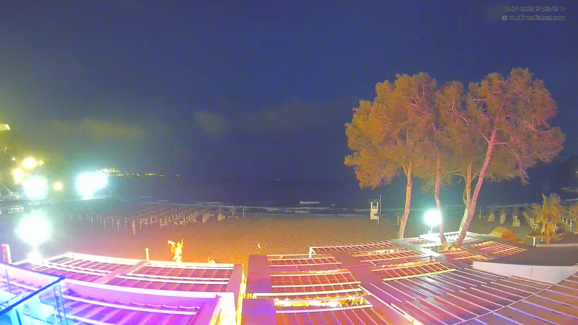 A nighttime view of a beach with a calm sea, several illuminated beach umbrellas, and trees near a beachfront structure with pink lighting.