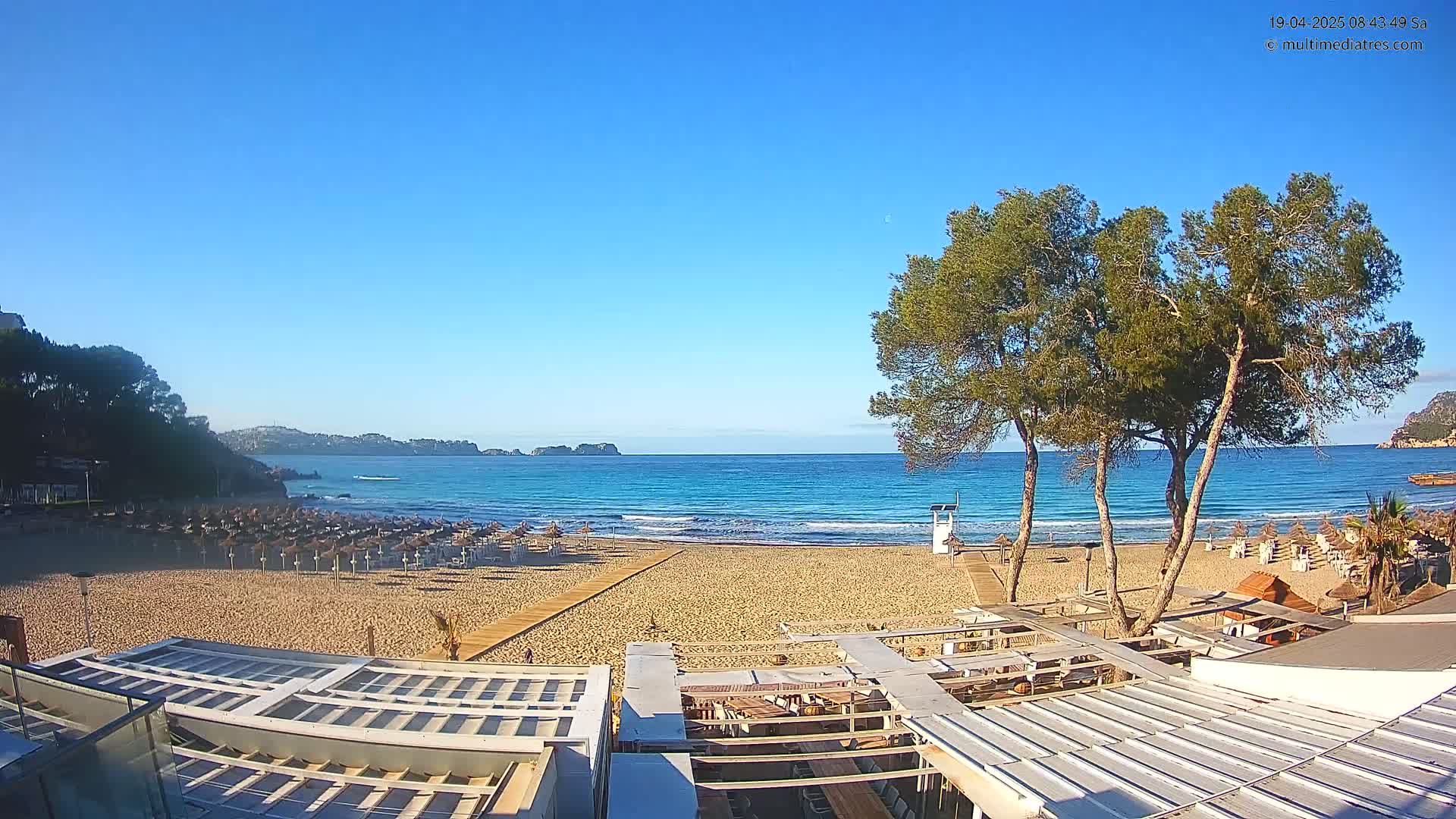 A sandy beach with rows of closed umbrellas stretches to a calm, turquoise ocean under a clear blue sky.