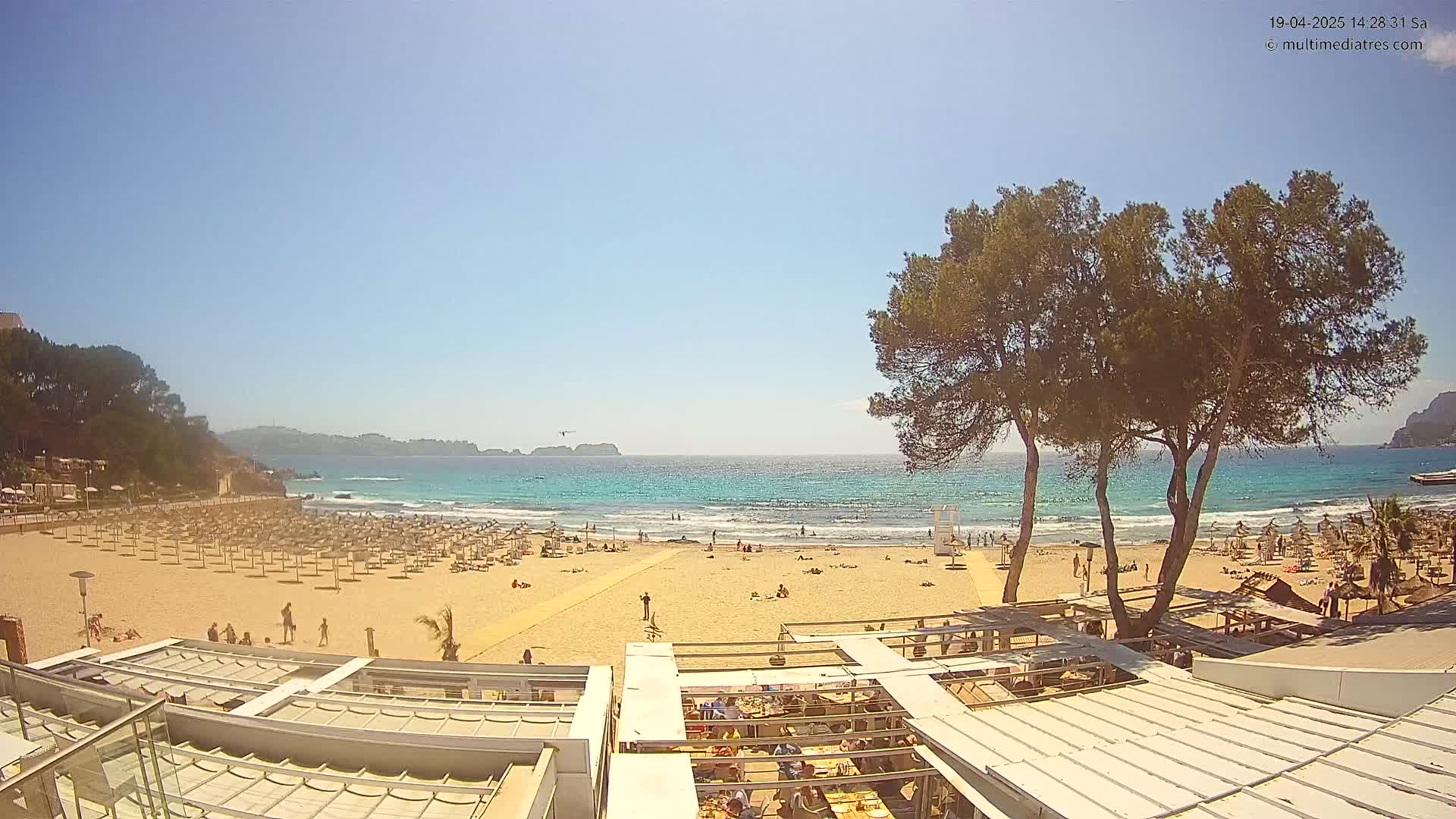 A sandy beach with numerous umbrellas and people is shown, overlooking a calm turquoise ocean under a clear blue sky.