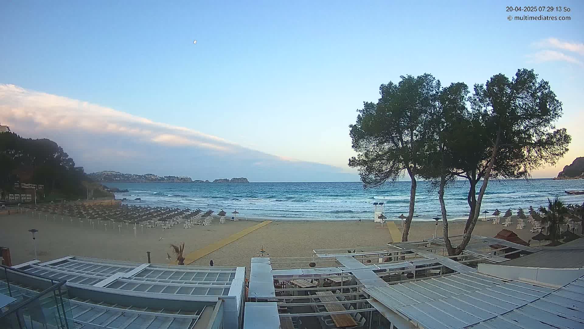 A mostly clear, twilight sky overlooks a sandy beach with rows of closed umbrellas, a calm sea with small waves, and trees near a beachfront building.