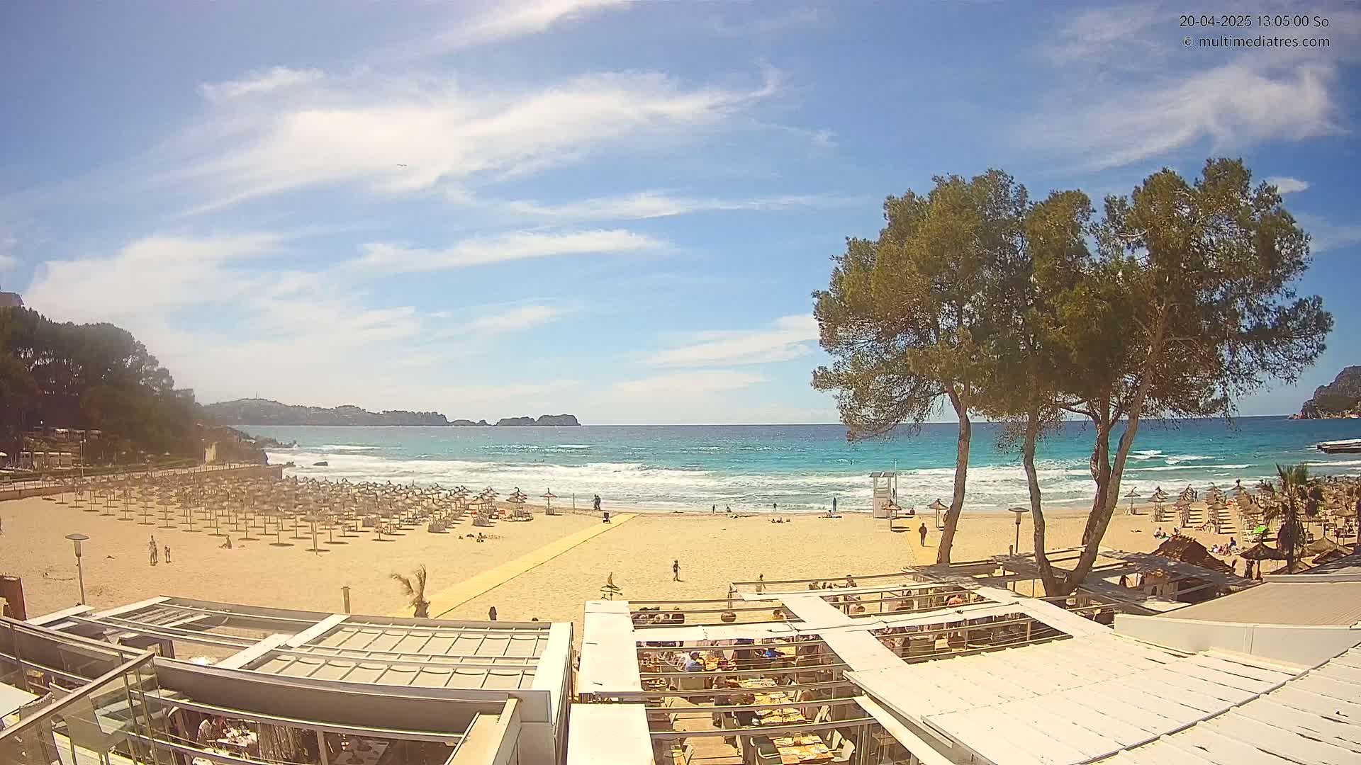 A sandy beach with numerous umbrellas and beachgoers is visible beneath a mostly sunny sky, overlooking a calm ocean with small waves.
