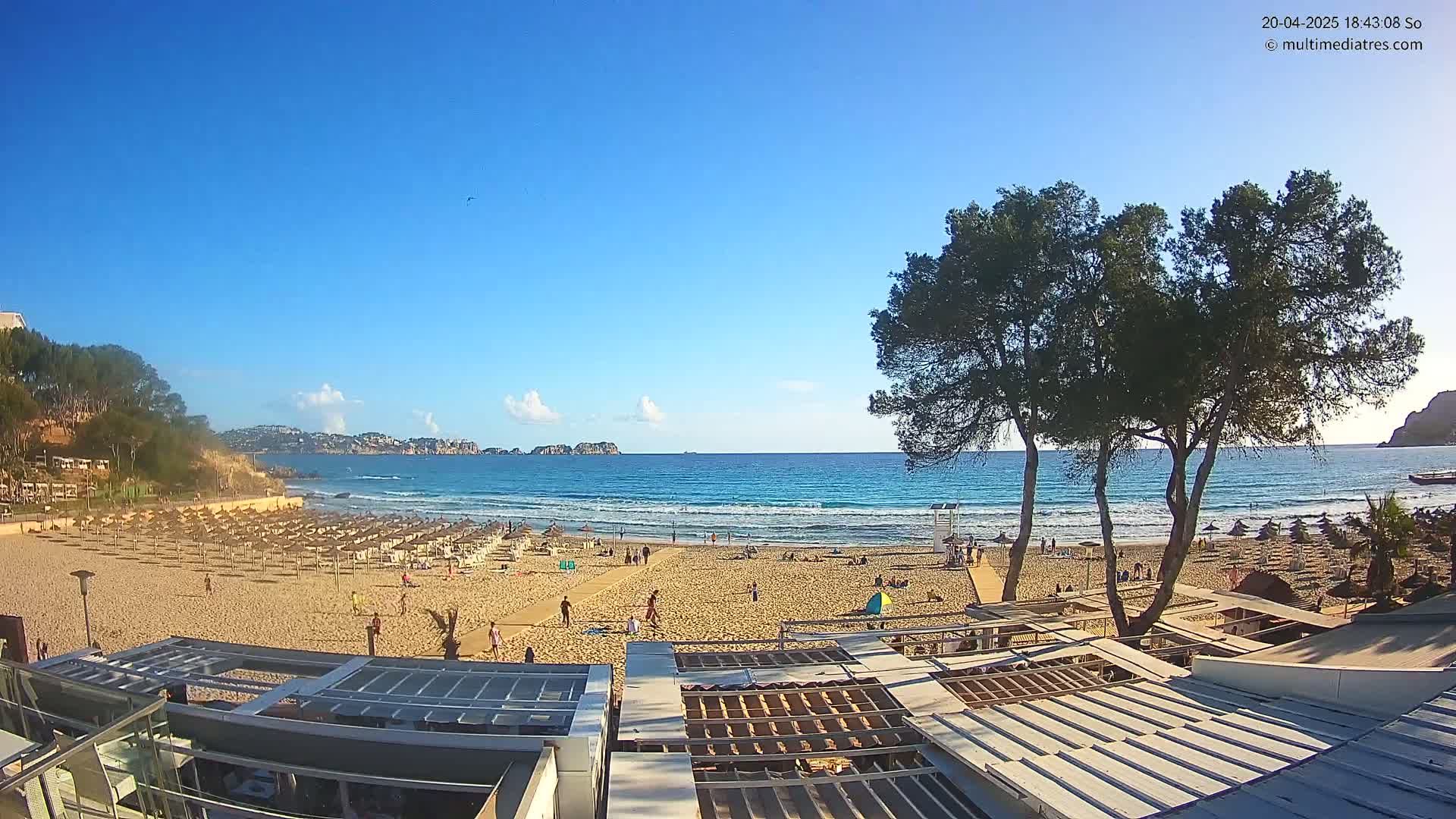 A sandy beach with many umbrellas and scattered people is seen under a clear blue sky, overlooking a calm ocean with small waves.