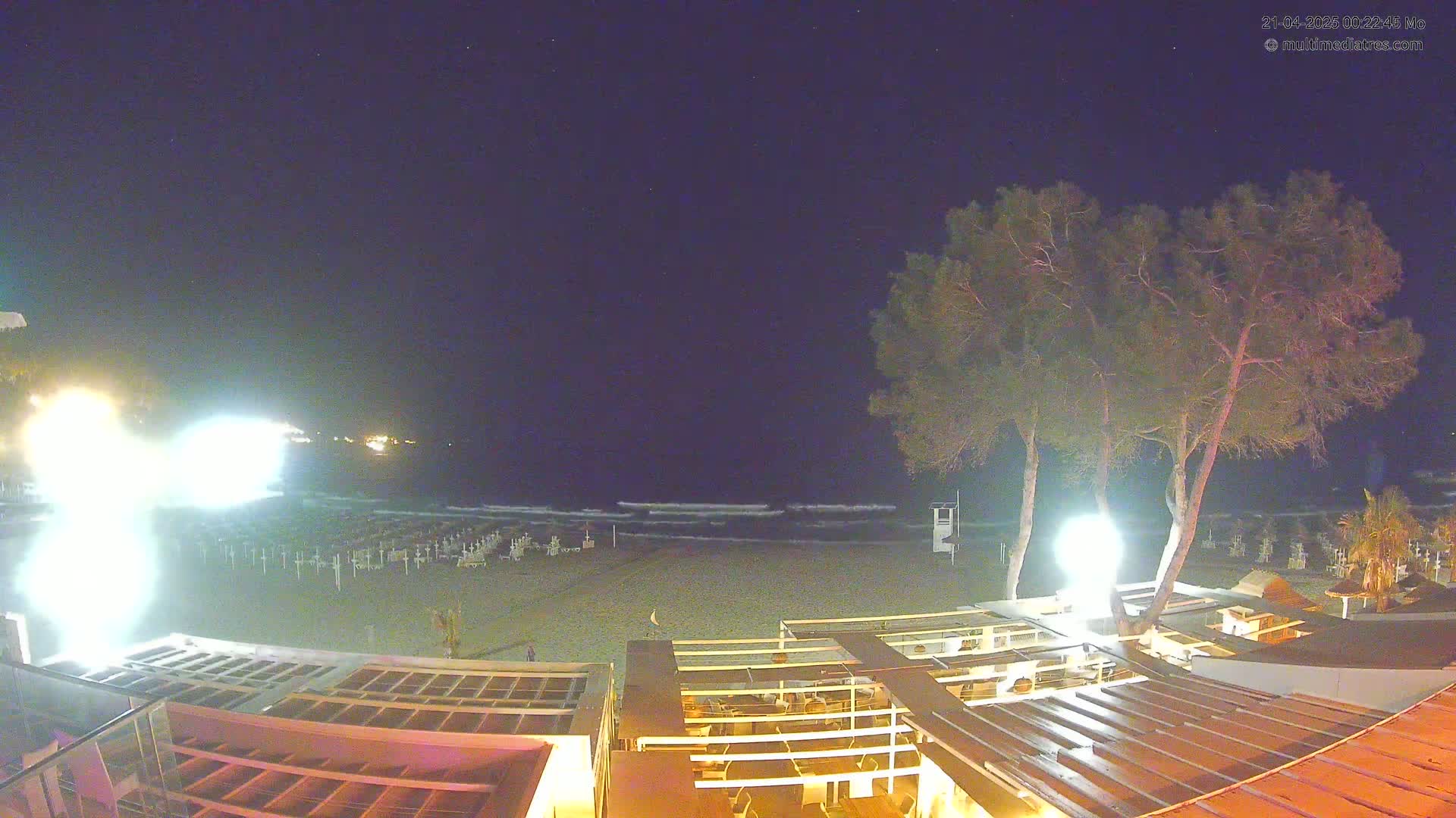 A nighttime beach scene shows a sandy shore with gently lapping waves under a clear, dark sky, illuminated by artificial lights near the beachside structures.