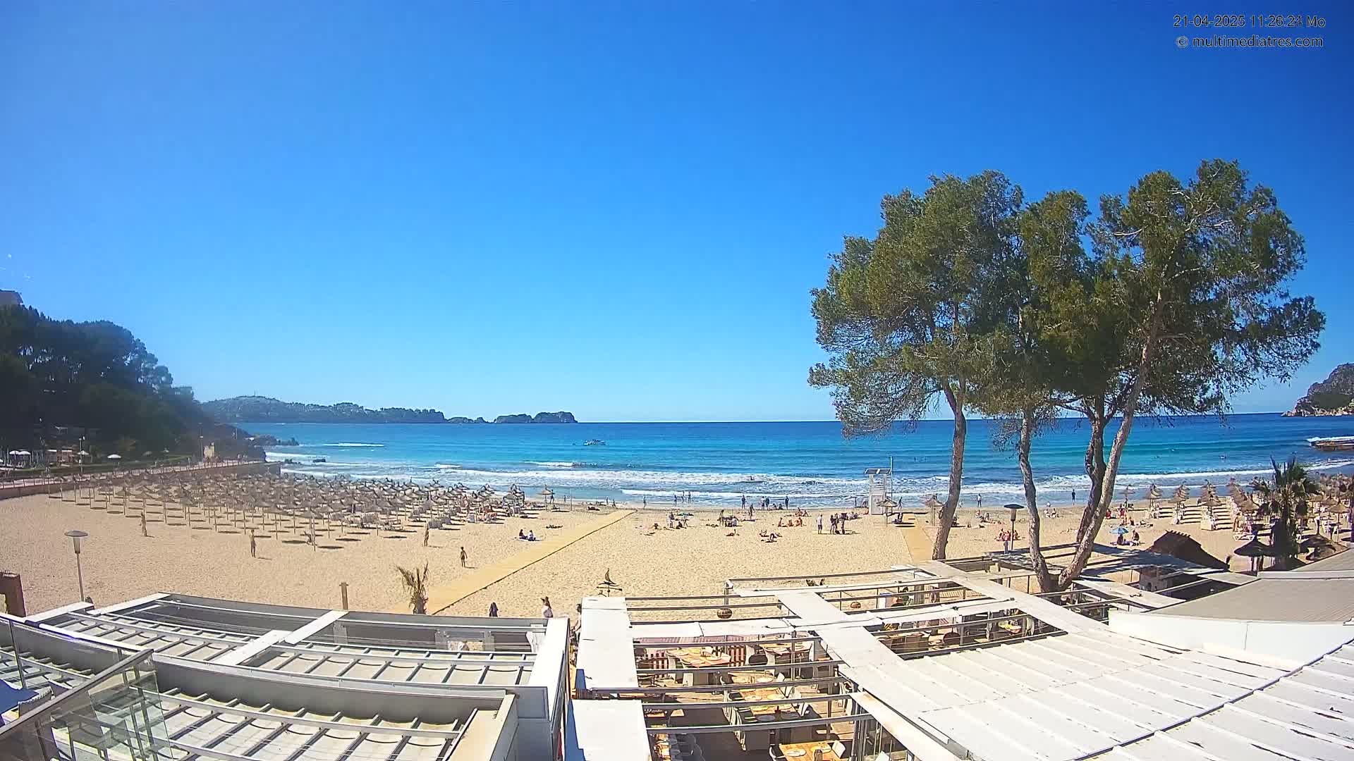 A sandy beach with numerous umbrellas and people enjoying a sunny day, overlooks a calm ocean with small waves under a clear blue sky.